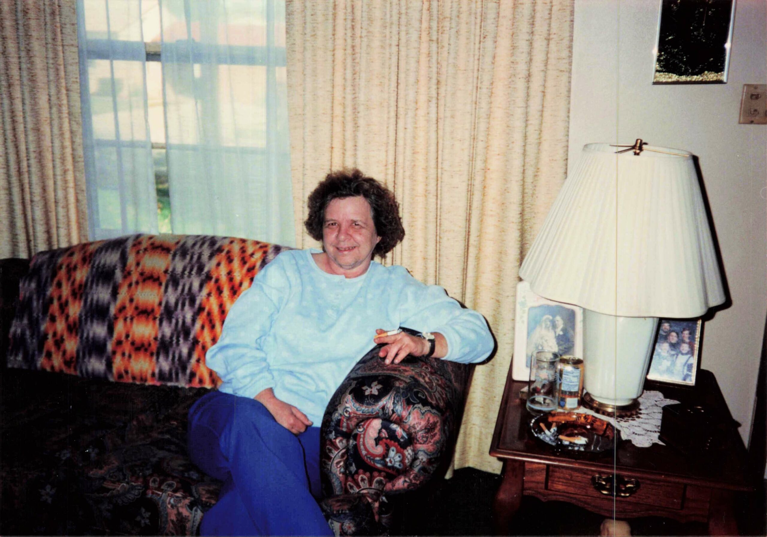 Older woman seated on a patterned couch in a living room with beige curtains, smiling at the camera.