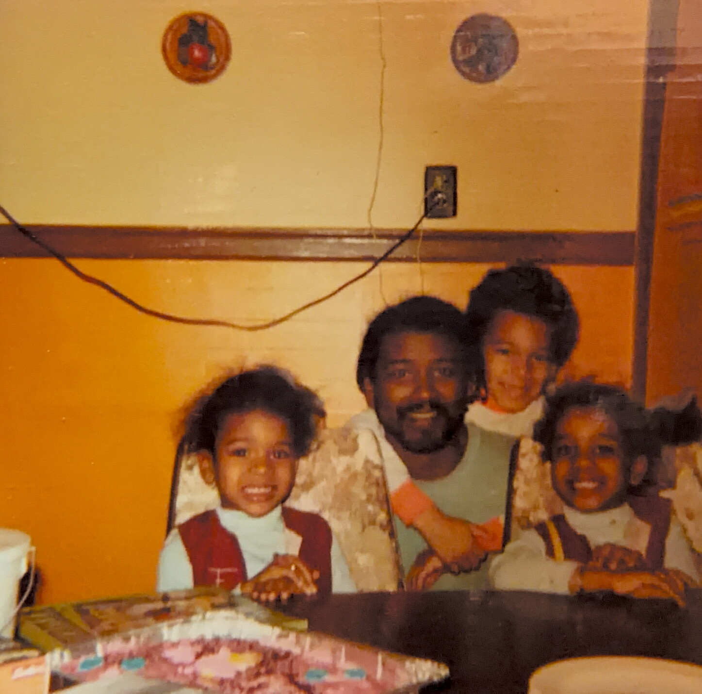 Family of four smiling for a birthday photo, sitting at a table with a cake and presents in front.