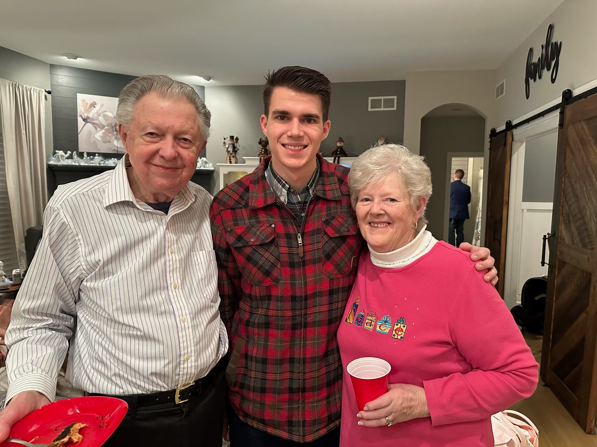 Three smiling people pose for a photo at a holiday gathering, each holding a red cup inside a warmly lit living room.