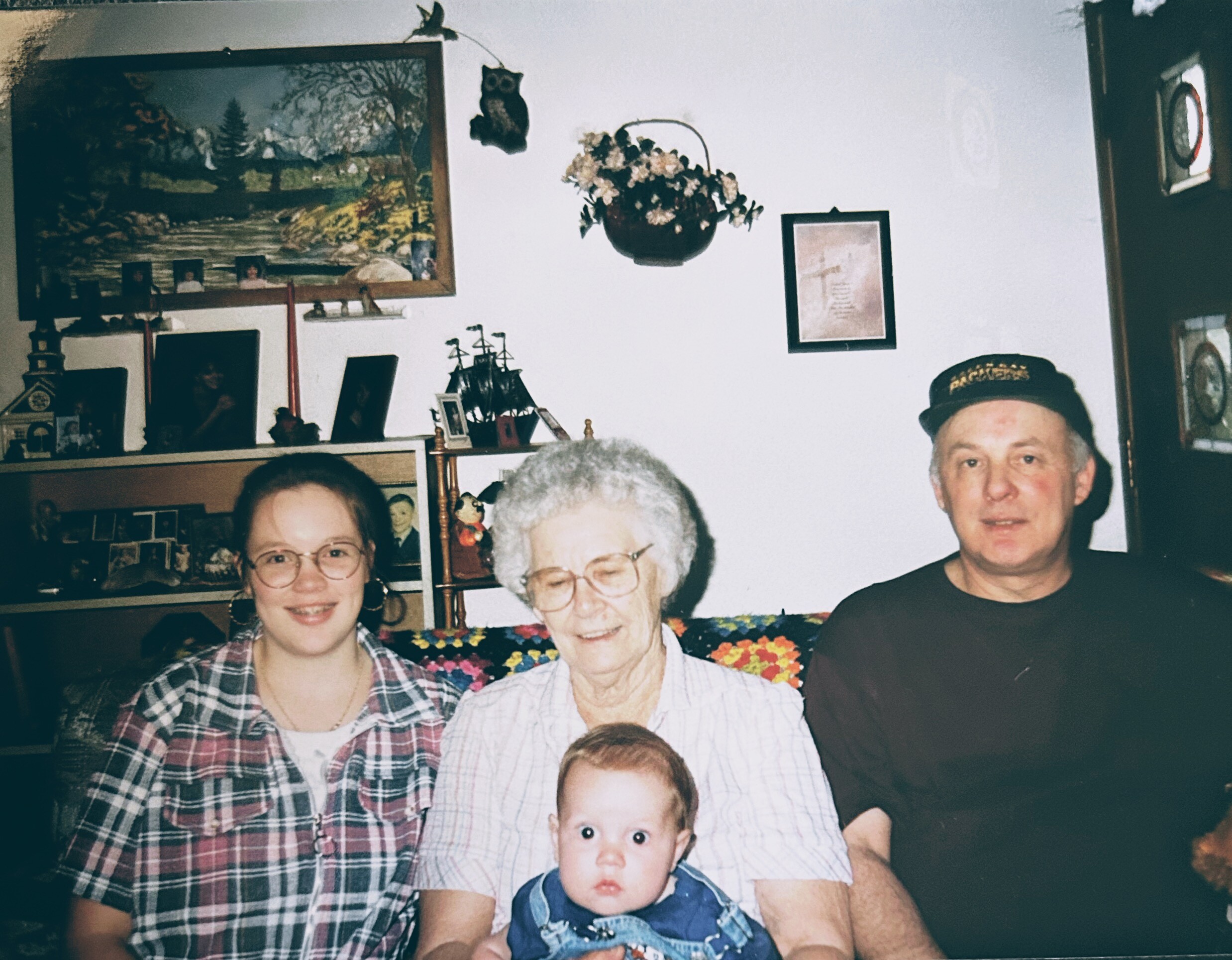 Group portrait of several people in a living room, including an elderly woman holding a baby, a man on the right, and a younger woman on the left, all smiling at the camera.