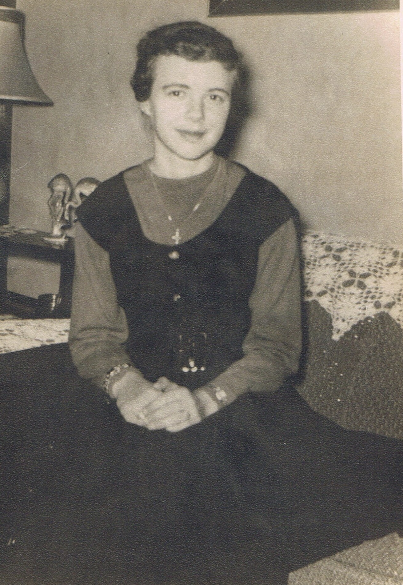 Woman sitting indoors with hands folded in her lap, wearing a dark sleeveless top over a long-sleeve shirt and a necklace.