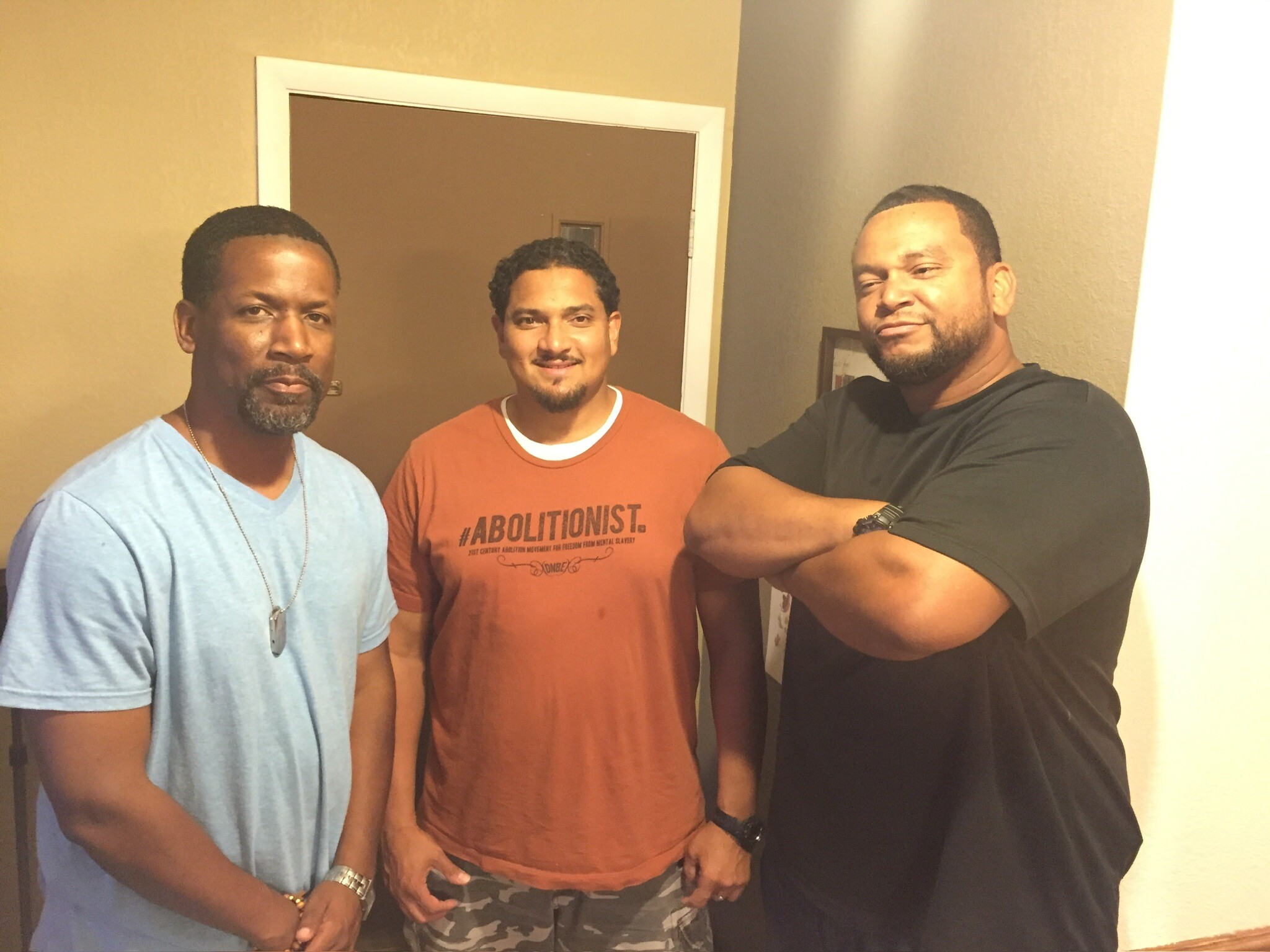 Three men posing together for a photo in a doorway, casual shirts (blue, orange, black) and relaxed smiles.