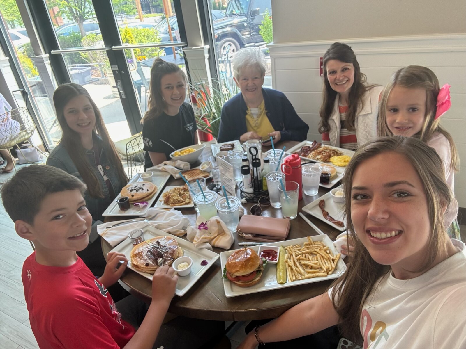 Group of kids and an elderly woman seated around a round table in a bright cafe, smiling with plates of pancakes, burgers, and fries in front of them.
