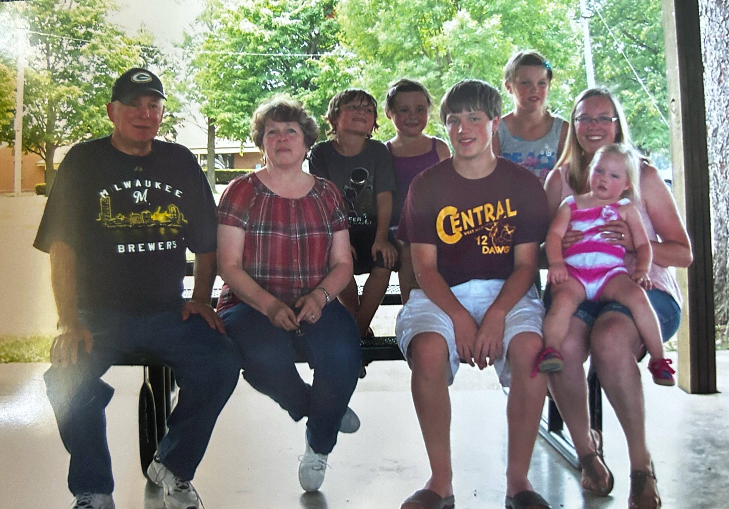 Group portrait of a multi-generation family sitting on a bench indoors/outdoors, smiling at the camera, with adults and children including a baby.