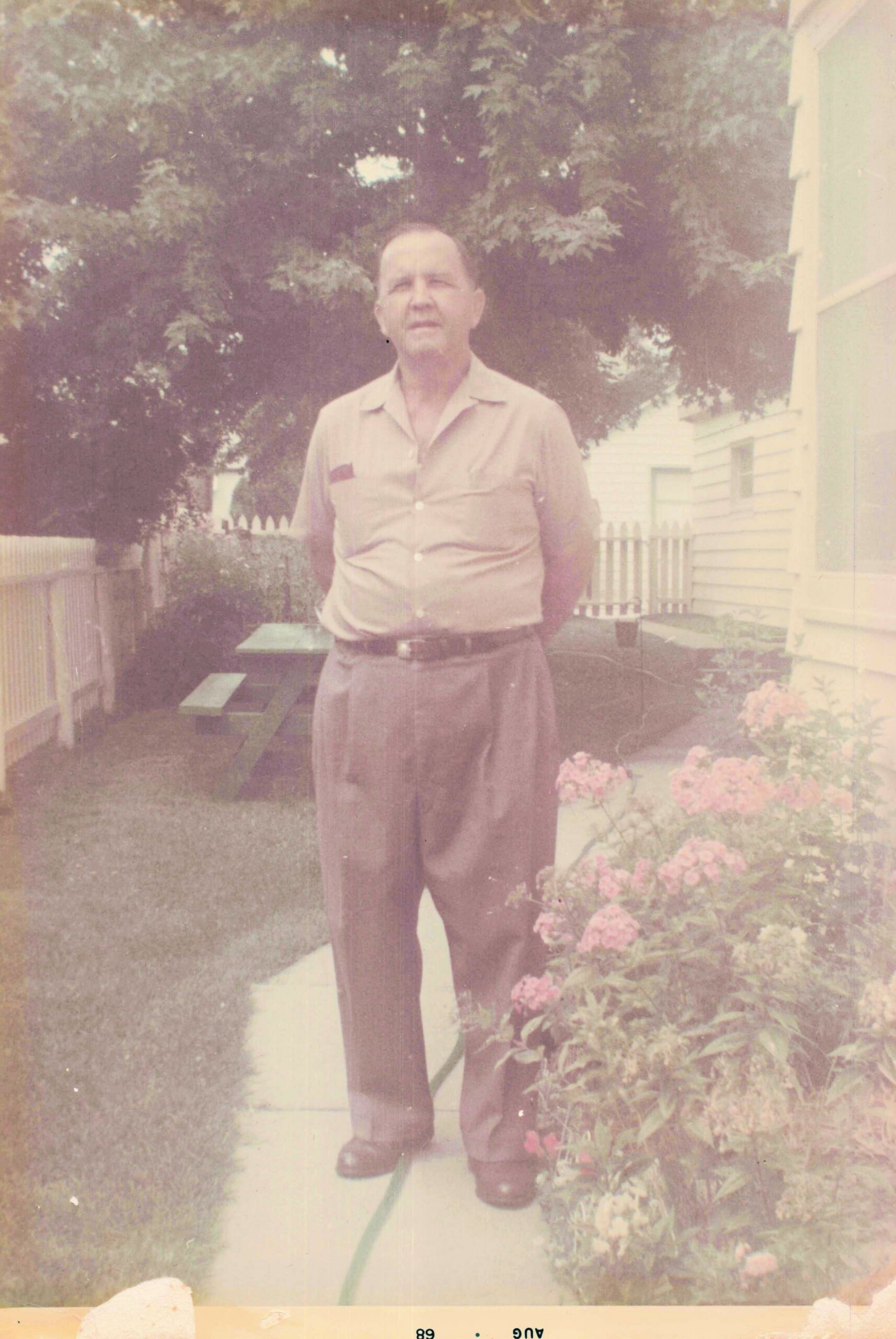Older man posing in a backyard garden, wearing a light button-down shirt, belt, and high-waisted pants, with a fence and house in the background.