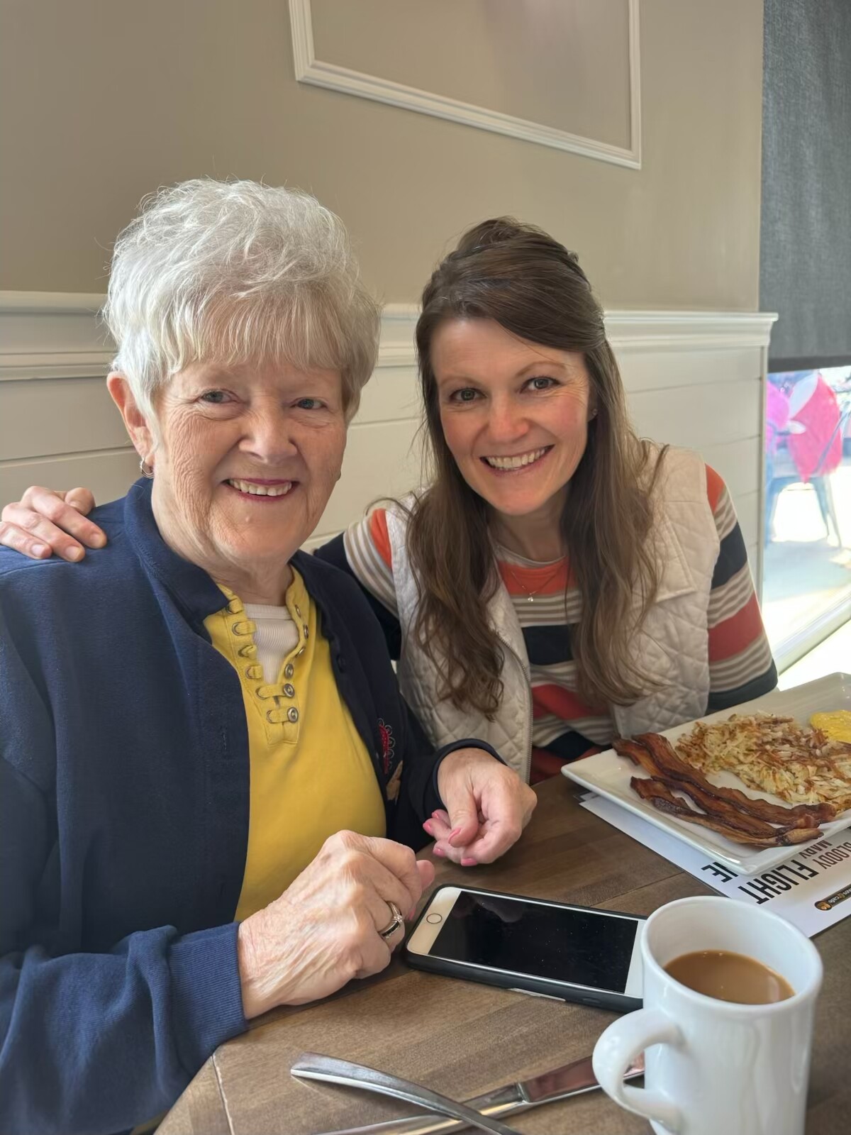 Two smiling women sit at a café table together, one elderly with white hair and the other younger, with a plate of bacon and eggs in front of them.