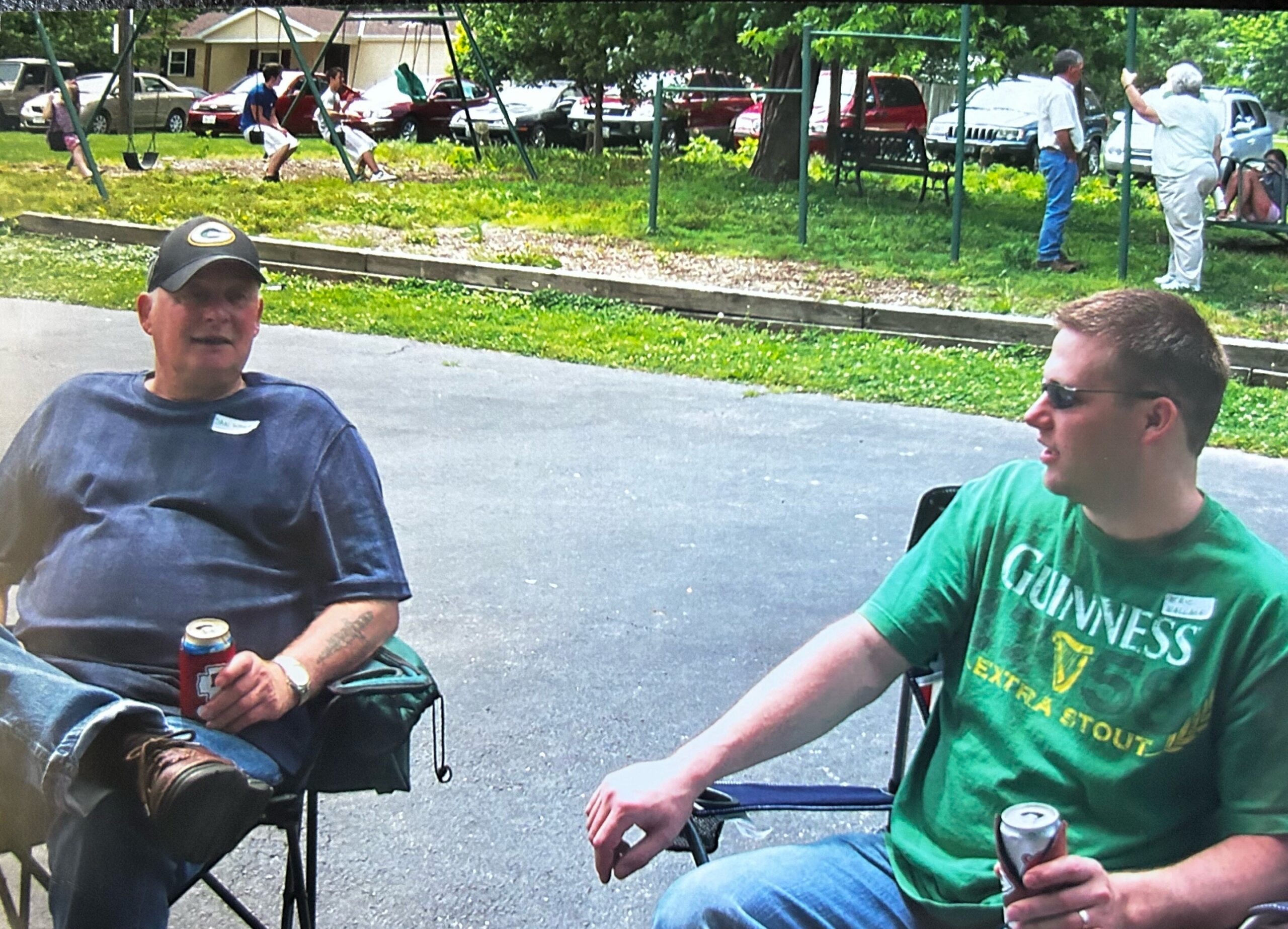 Two men sit in lawn chairs outdoors, holding cans, with a park and parked cars behind them in a casual gathering.