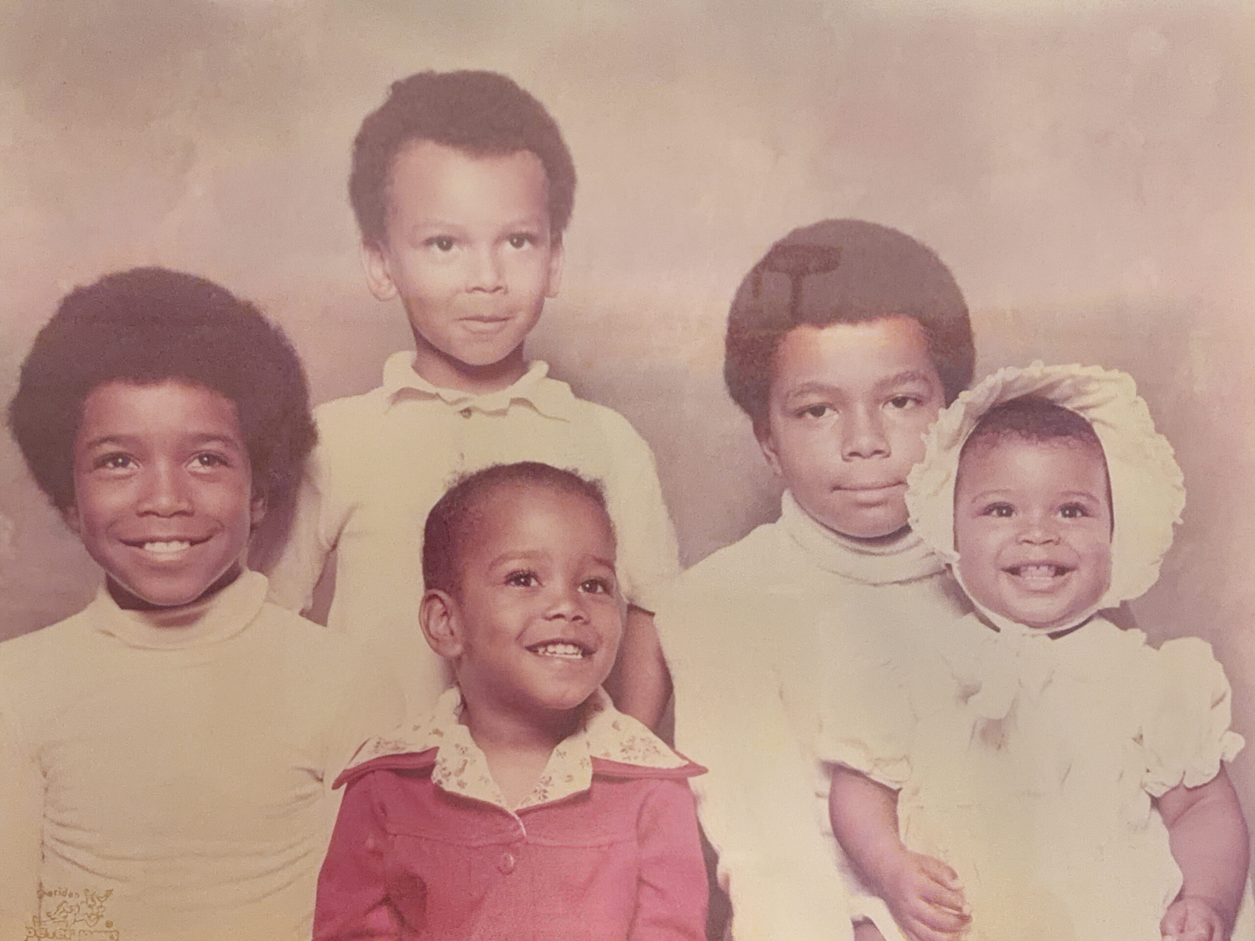 Group of smiling children in a faded vintage studio portrait, wearing light-colored clothing against a beige backdrop.