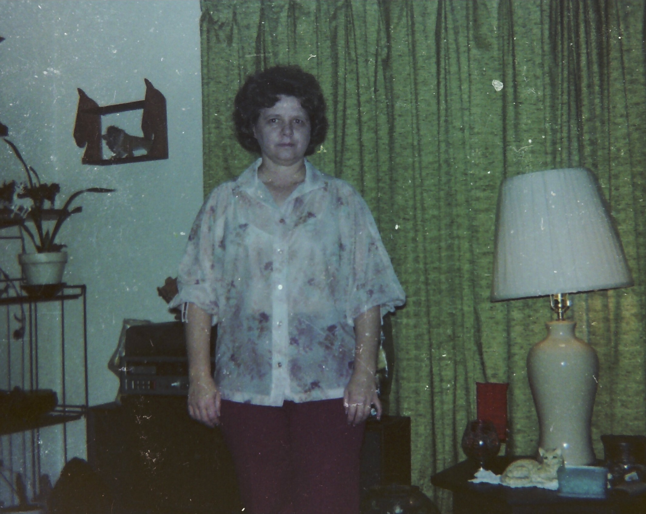 Woman standing in a living room wearing a light floral blouse, with green striped curtains and a lamp in view behind her.