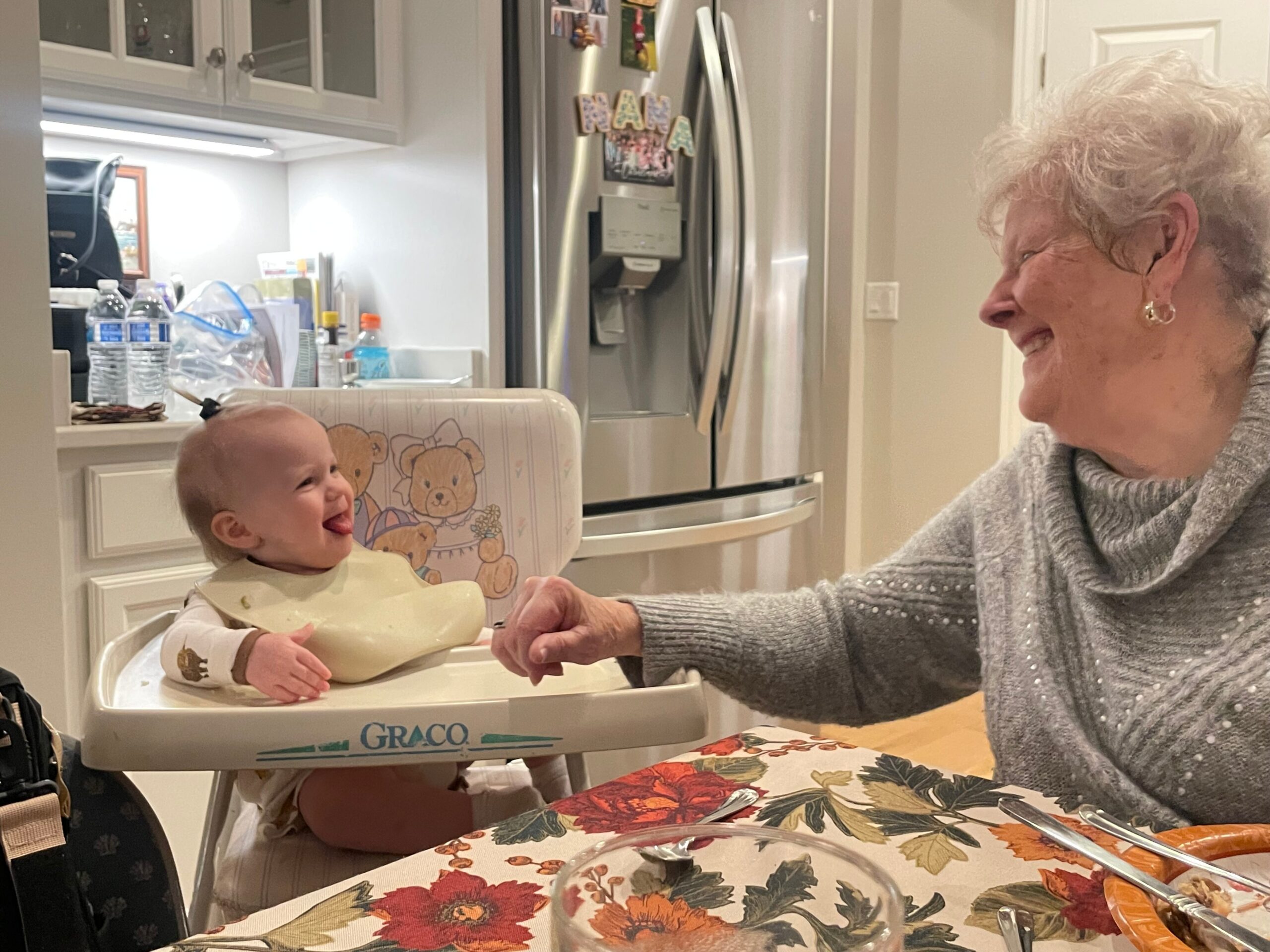 Baby in a Graco high chair laughing as an elderly woman smiles and reaches toward him in a bright kitchen environment.