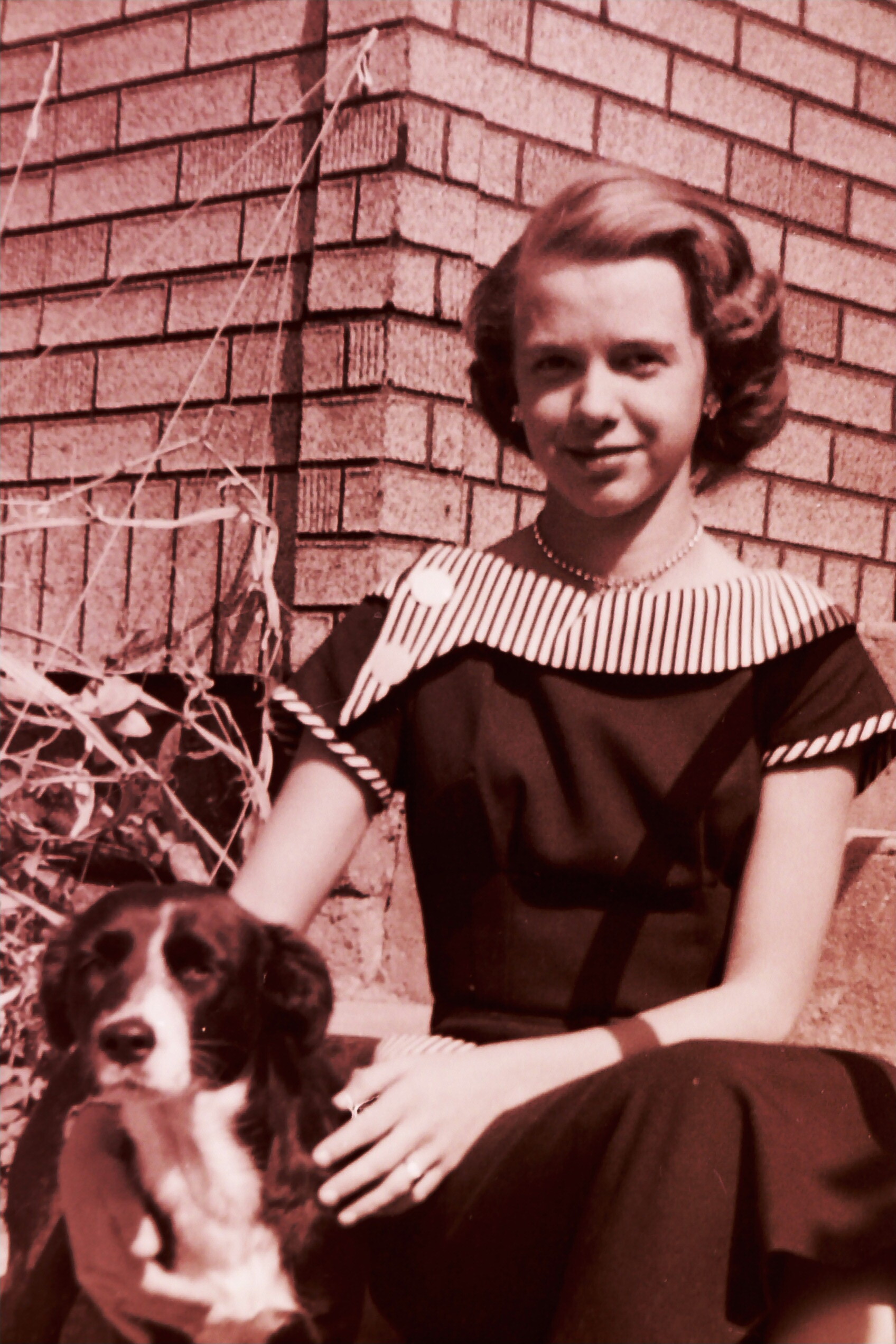 Young woman in a vintage dress with a striped collar sits beside a black-and-white dog against a brick wall outdoors.