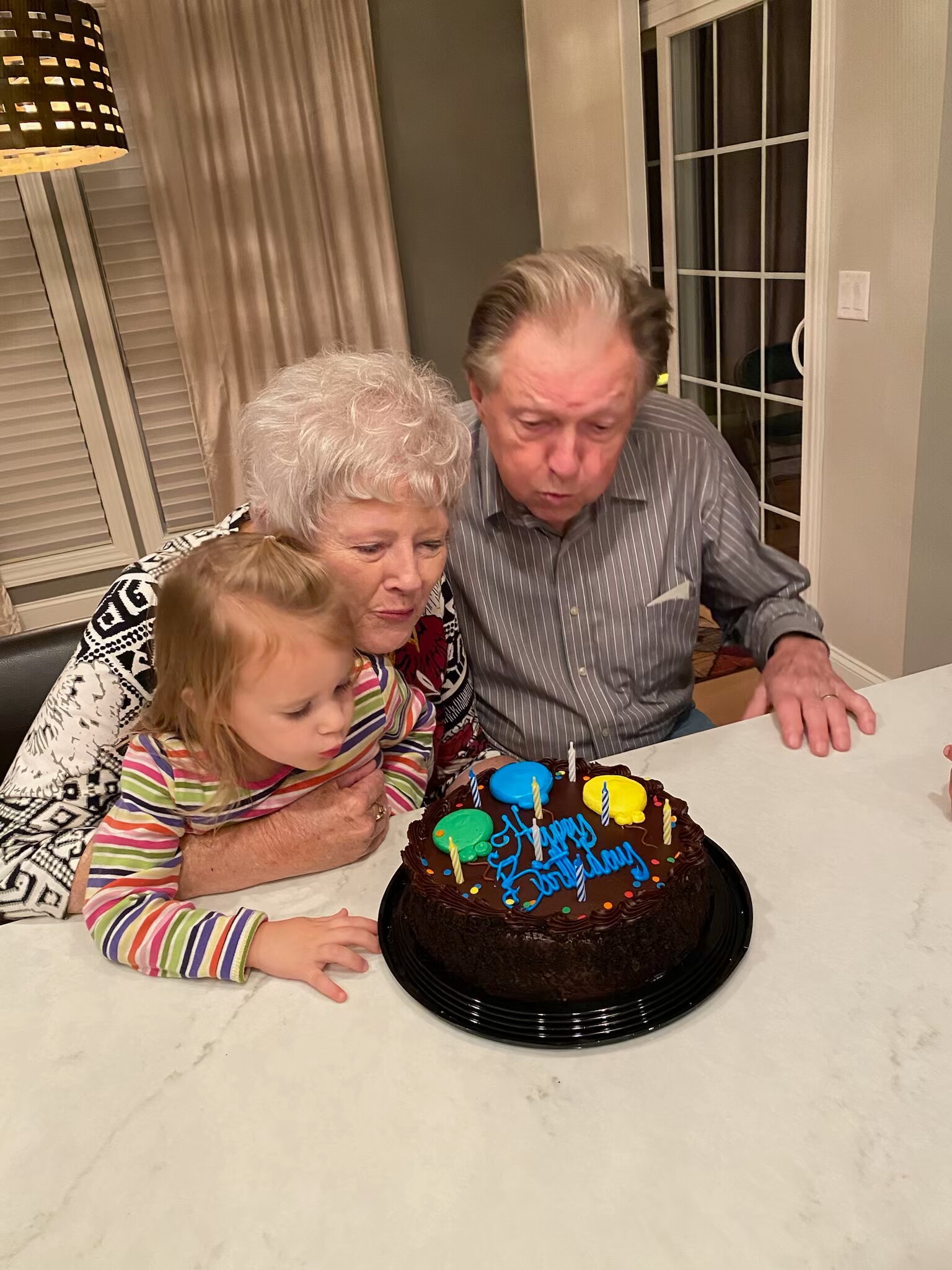 Grandparents and a young girl lean in to celebrate a chocolate birthday cake with candles at a kitchen table.