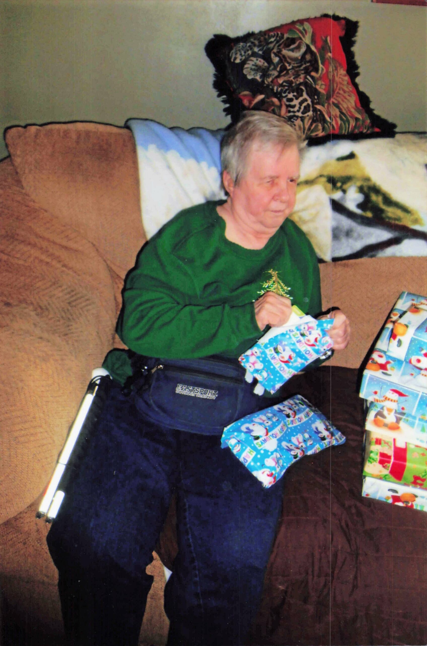Older person in a green sweatshirt sits on a brown couch, opening colorful wrapped gifts for a celebration.