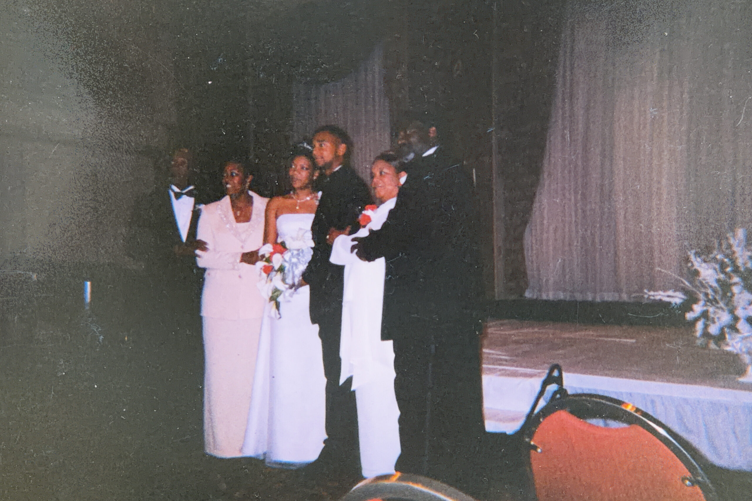 Bride in a white dress holds a bouquet, standing with the groom in a tuxedo and bridesmaids on a stage during a wedding pose on the left side of the image.