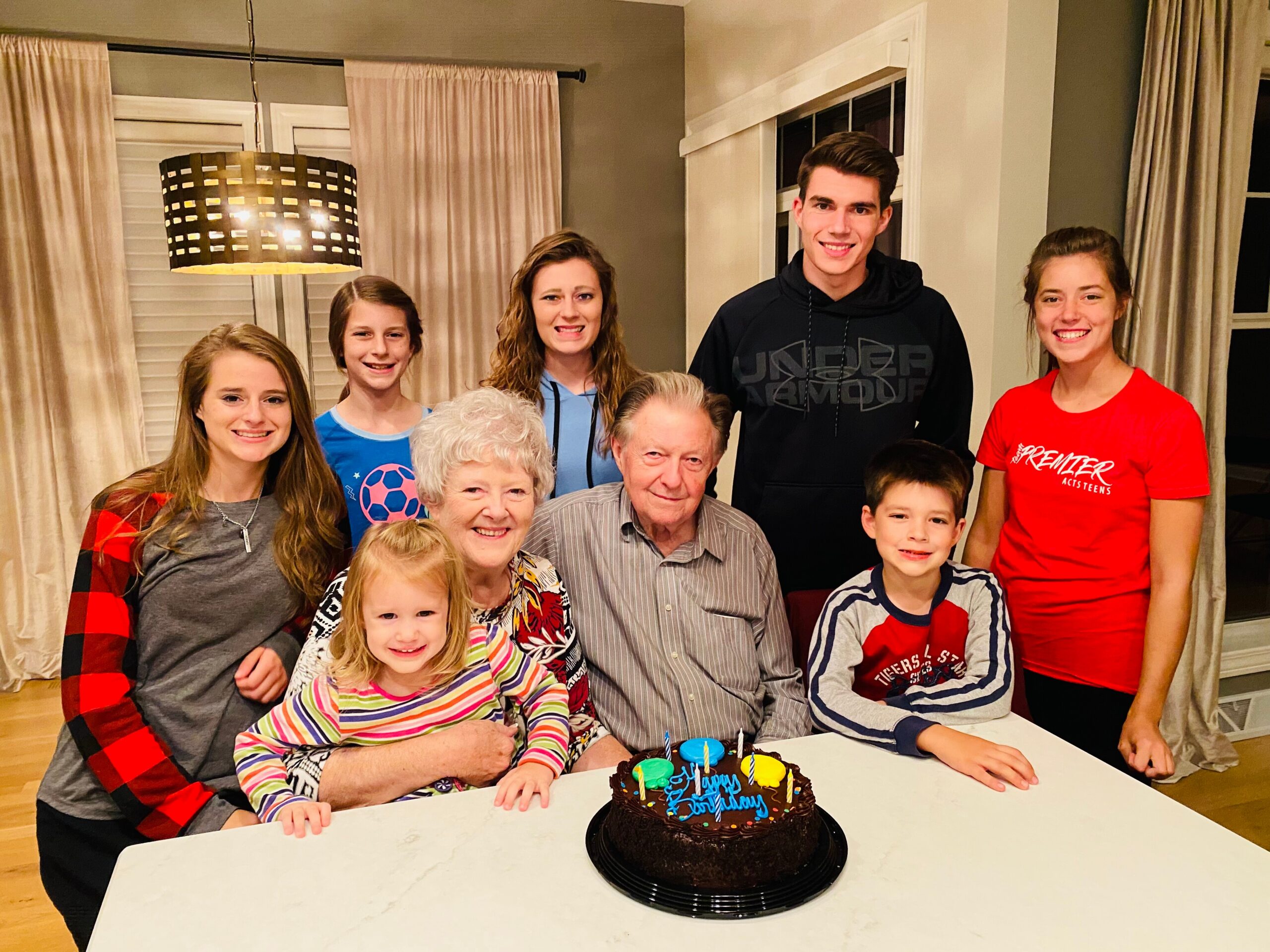 A group of ten family members gathered around a table with a chocolate birthday cake front and center, smiling for the photo.