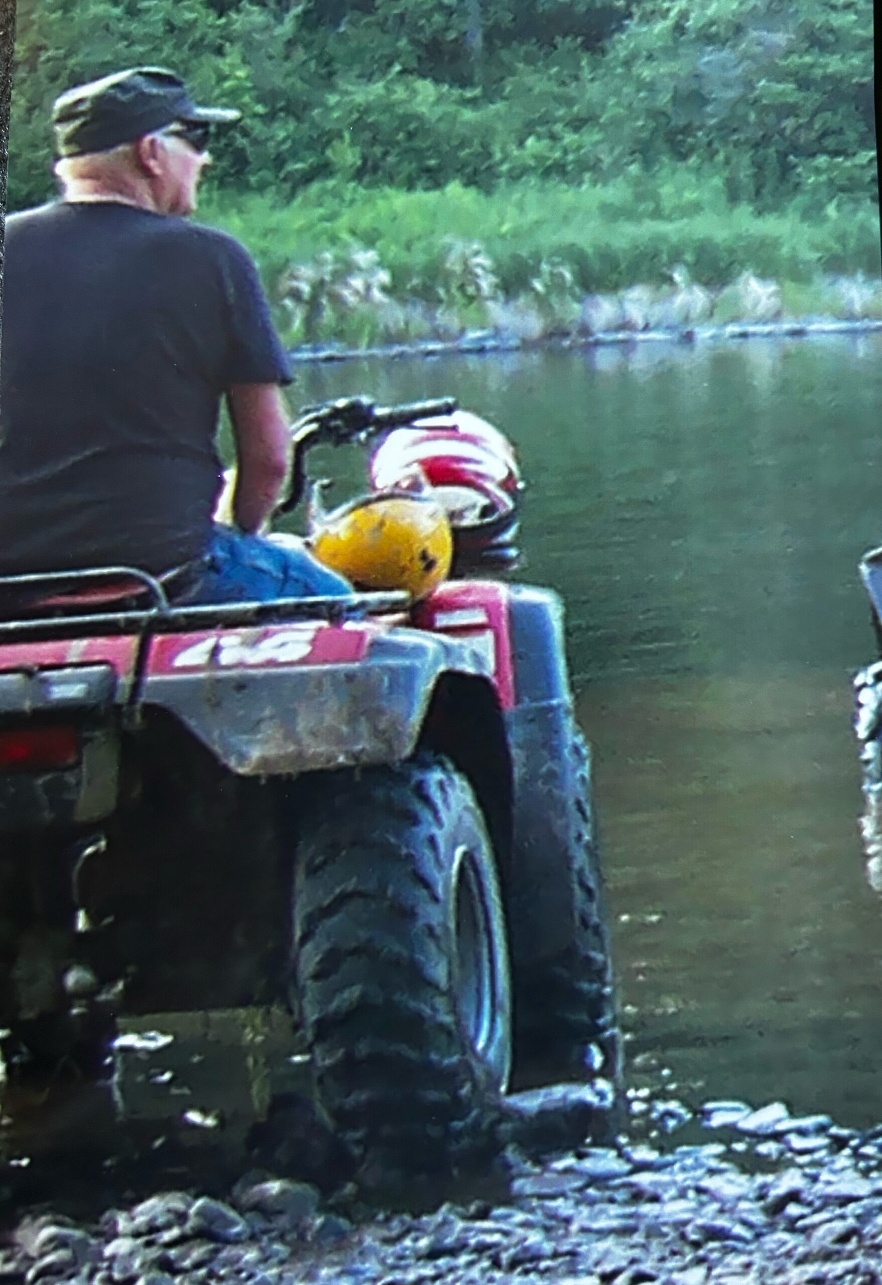Man seen from behind riding a red ATV along a rocky riverbank with dense green vegetation in the background.