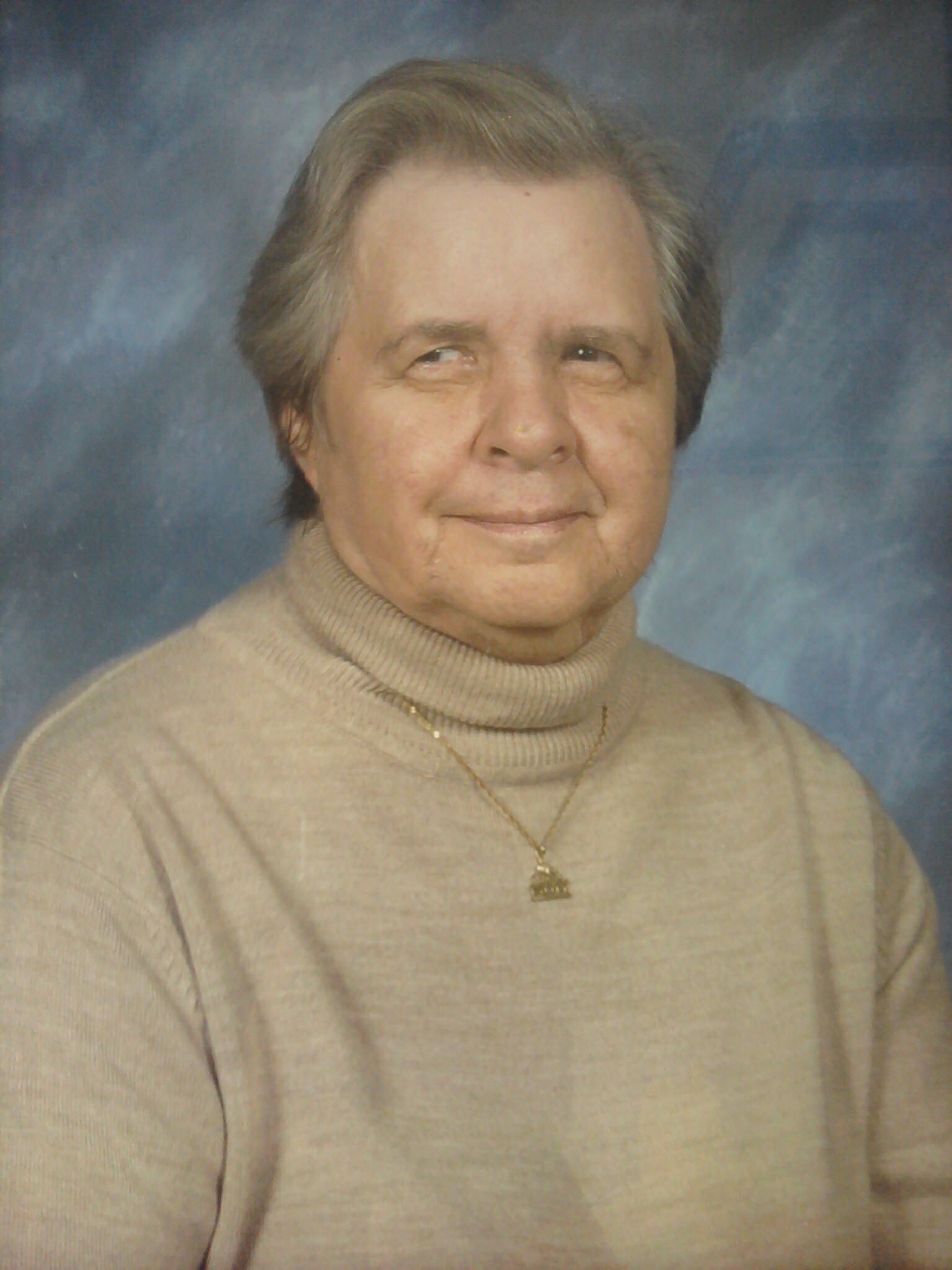 Portrait of an older woman wearing a beige turtleneck sweater and a gold pendant, with a blue studio backdrop behind her.