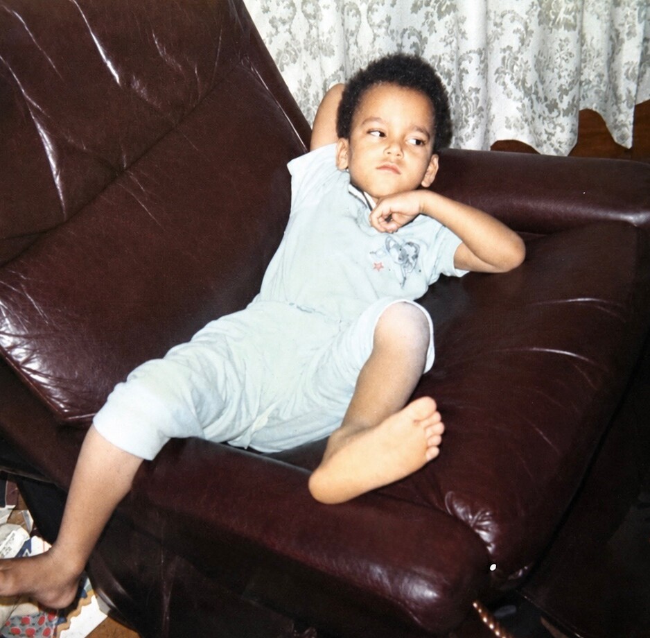 Young child lounging on a dark brown leather sofa with arm behind the head, relaxed pose.