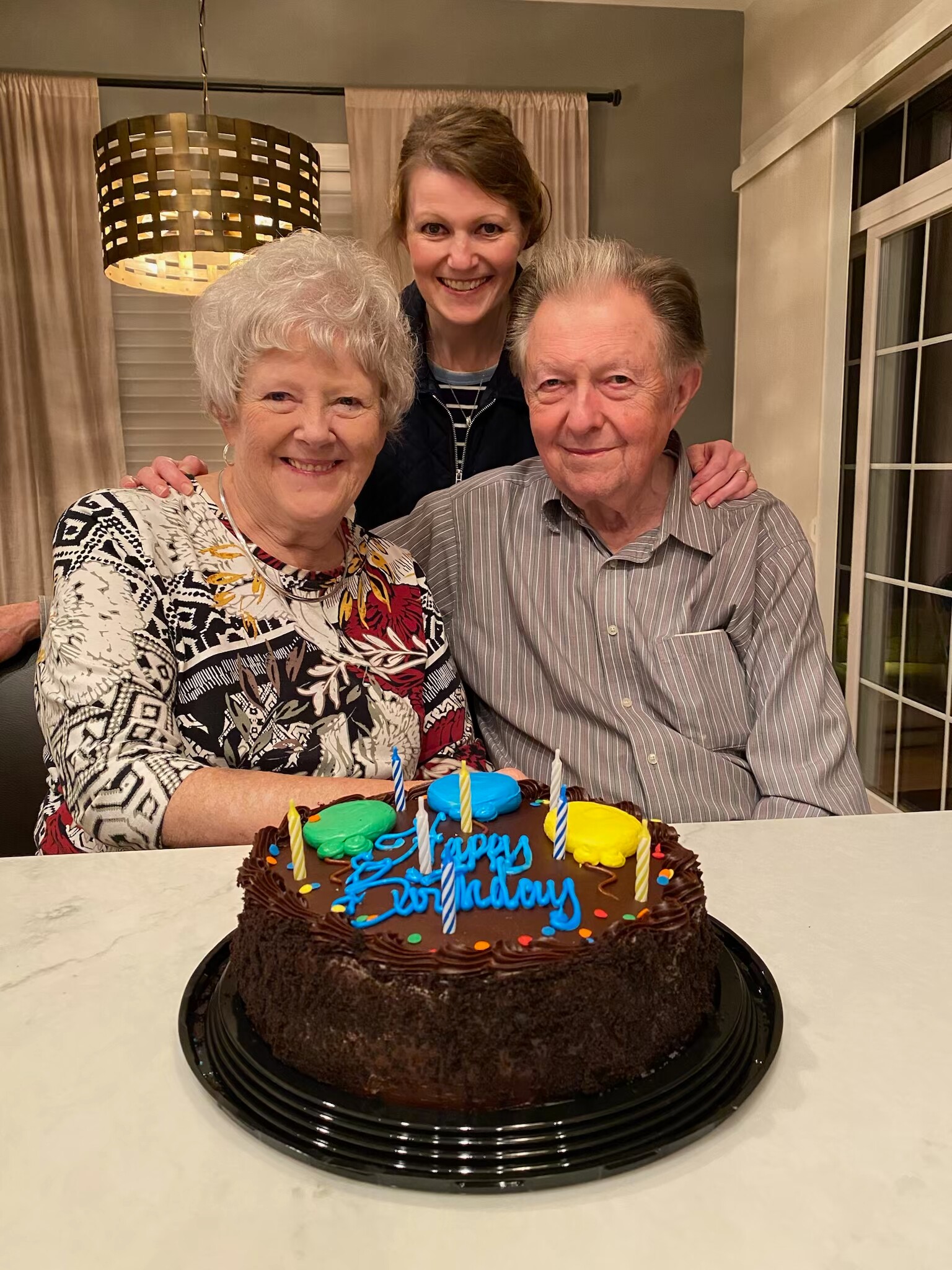 Elderly couple with a young woman behind them posing for a birthday photo by a chocolate cake with candles.