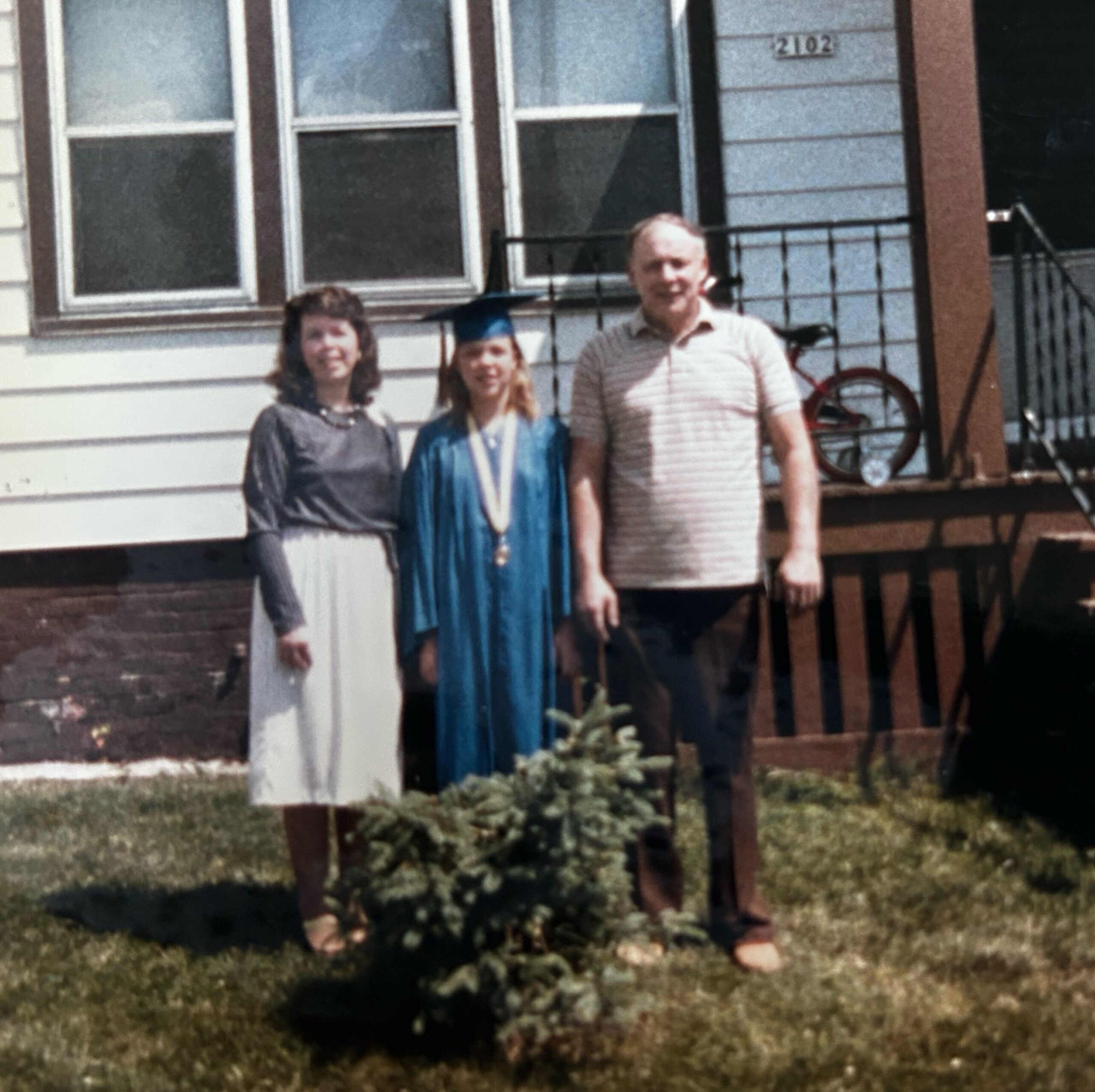 Three people posing on a lawn in front of a white house; the center person wears a blue graduation gown and cap.