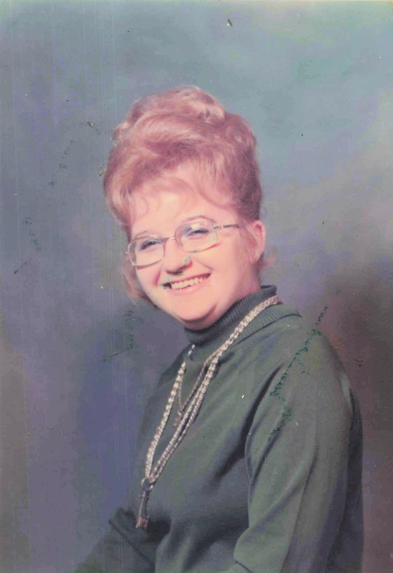 Smiling woman with short, styled hair and glasses, wearing a green turtleneck and a beaded necklace against a bluish backdrop.