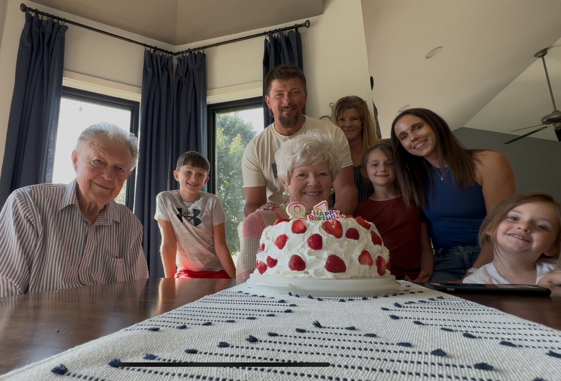 Eight family members pose around a large strawberry-topped birthday cake on a dining table, smiling for a photo in a bright home room.