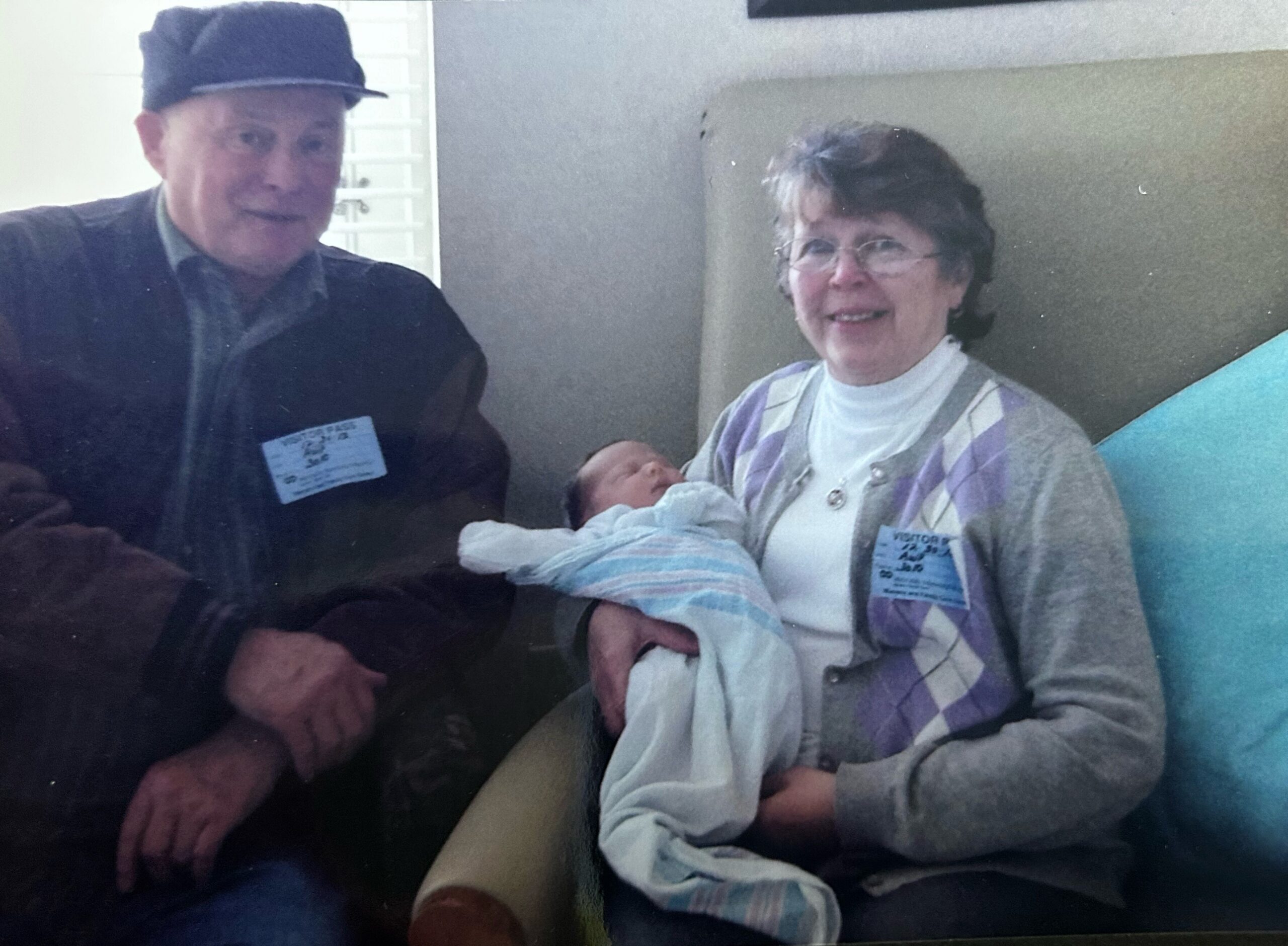 Older man wearing a dark cap sits beside a smiling woman holding a newborn wrapped in a striped blanket.