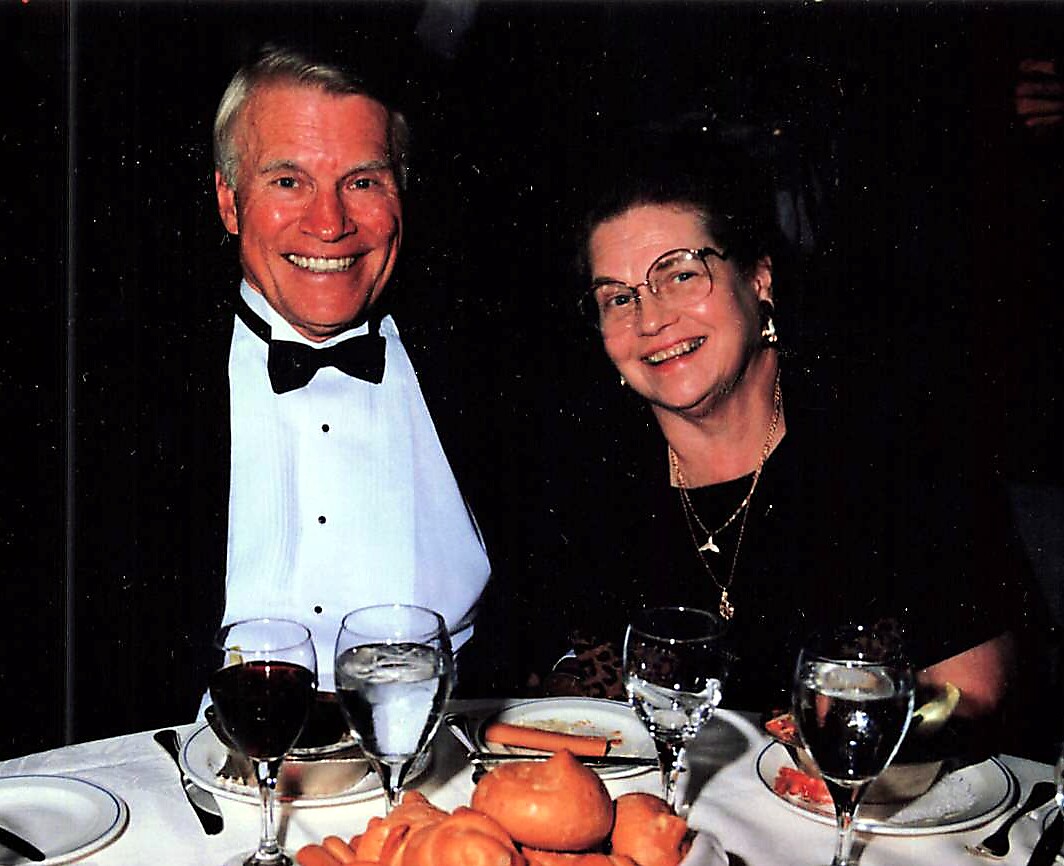 Smiling elderly couple in formal attire at a dinner table with lobster and wine glasses in front.