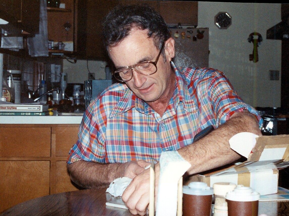 Man wearing a plaid shirt seals a cardboard box at a kitchen table with packing tape nearby and supplies around him.