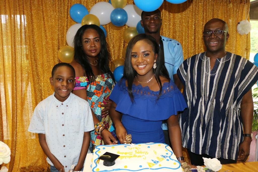 Six people gathered around a birthday cake with blue decorations in front of a gold sequin backdrop and balloons nearby.