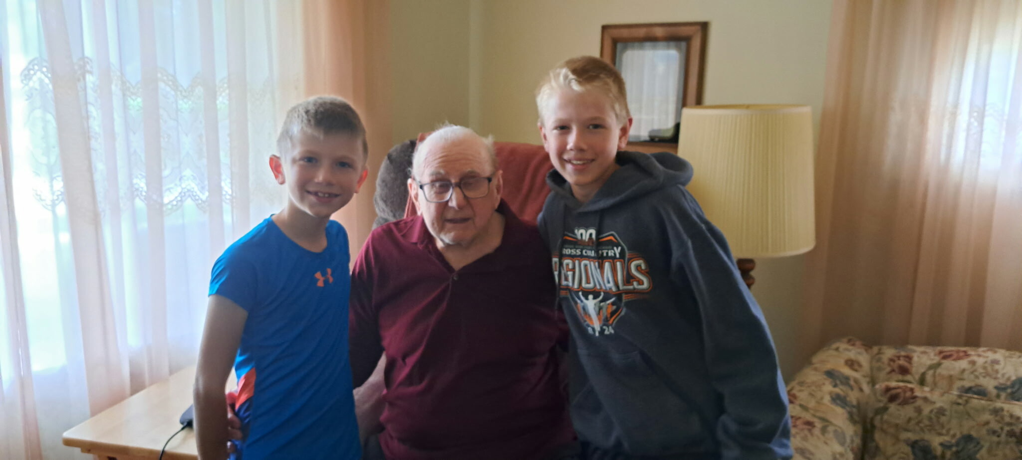 Three generations: an elderly man in a maroon polo flanked by two boys in blue and gray hoodies posing in a living room.