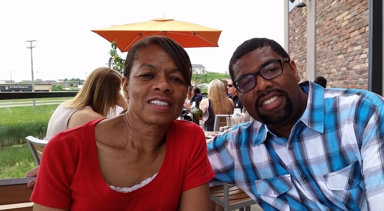 Two friends sit at an outdoor restaurant patio, smiling at the camera with an orange umbrella in the background.