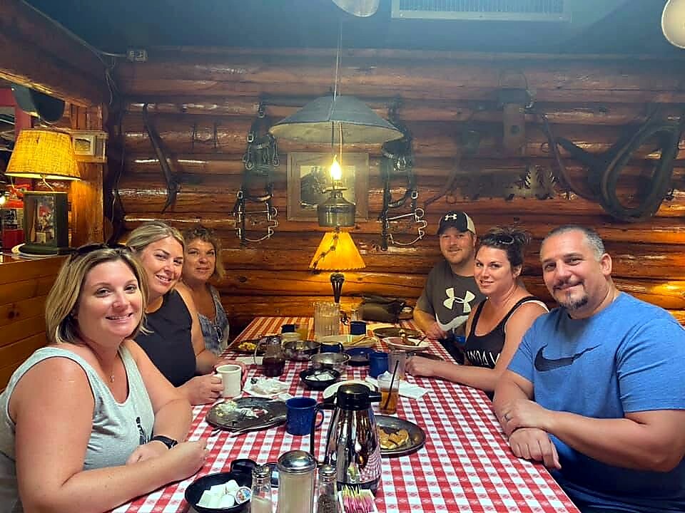 Six adults sit around a wood-paneled, rustic restaurant table with a red-and-white checkered tablecloth, smiling at the camera under a hanging lantern in a cozy setting.