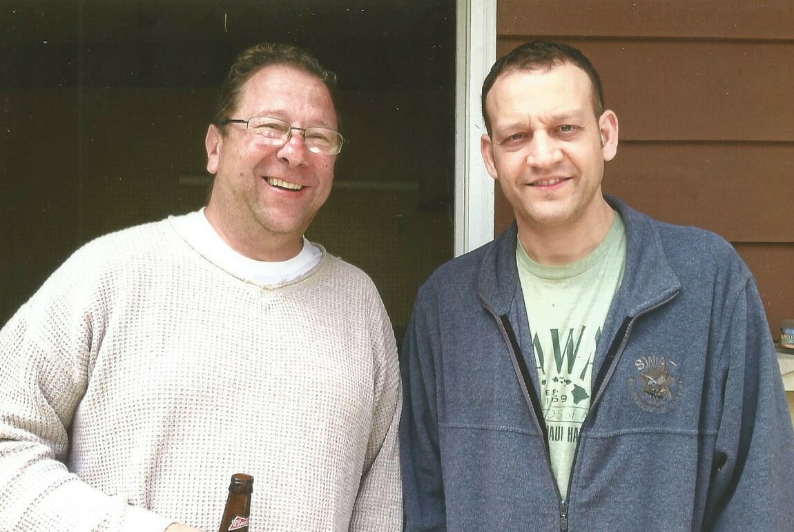 Two men smiling at the camera on a porch; one holds a beer bottle in his hand.