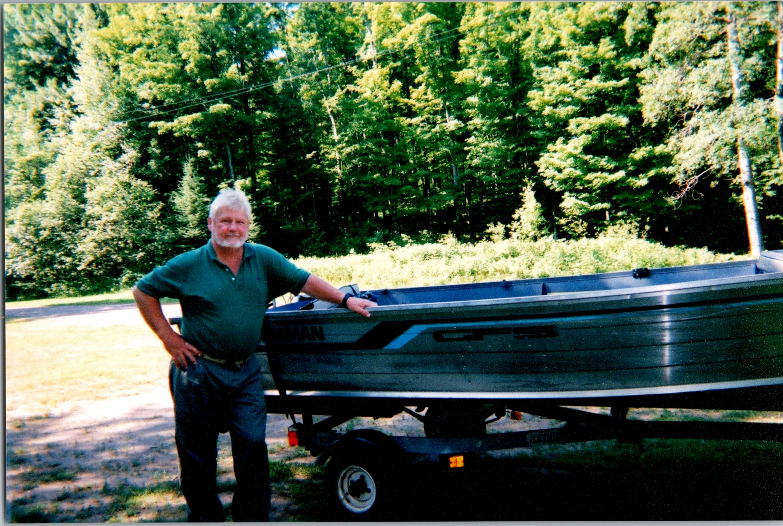 Man with gray beard standing beside a dark green boat on a trailer outdoors, with a forested background and sunshine.