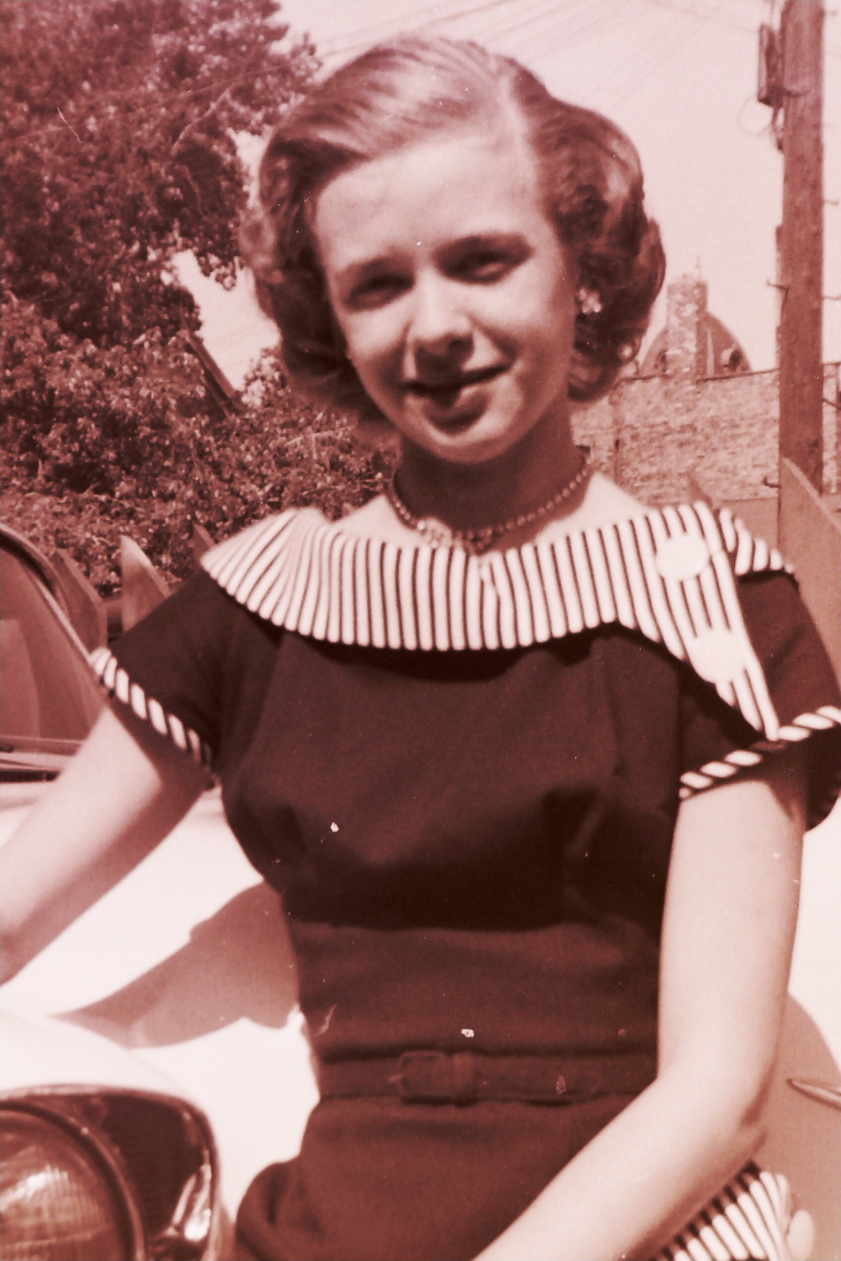 Young woman in a vintage dress with a striped collar, smiling outdoors near a car in a sepia-toned photo.