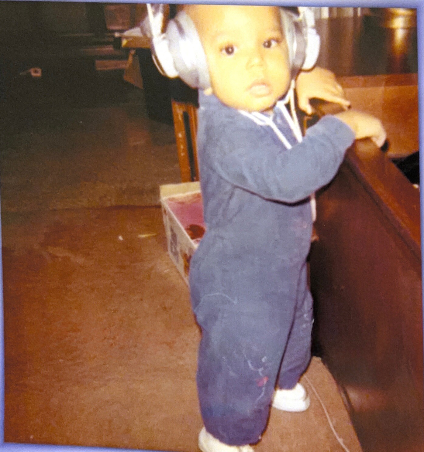 Toddler wearing oversized gray headphones and a blue onesie, standing indoors and gripping a wooden railing.
