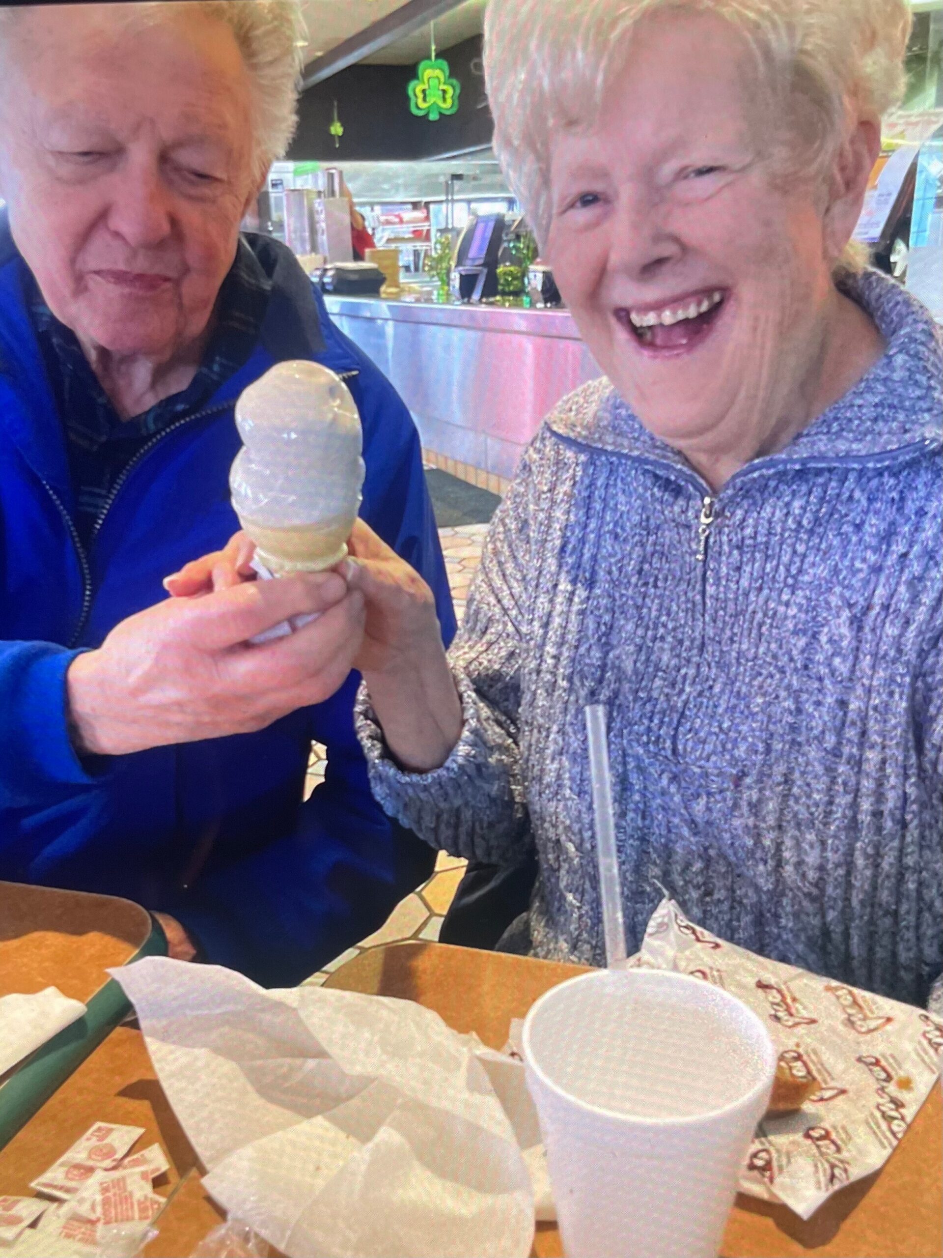 Two elderly friends share an ice cream cone at a cafe, one smiling broadly while holding the cone.
