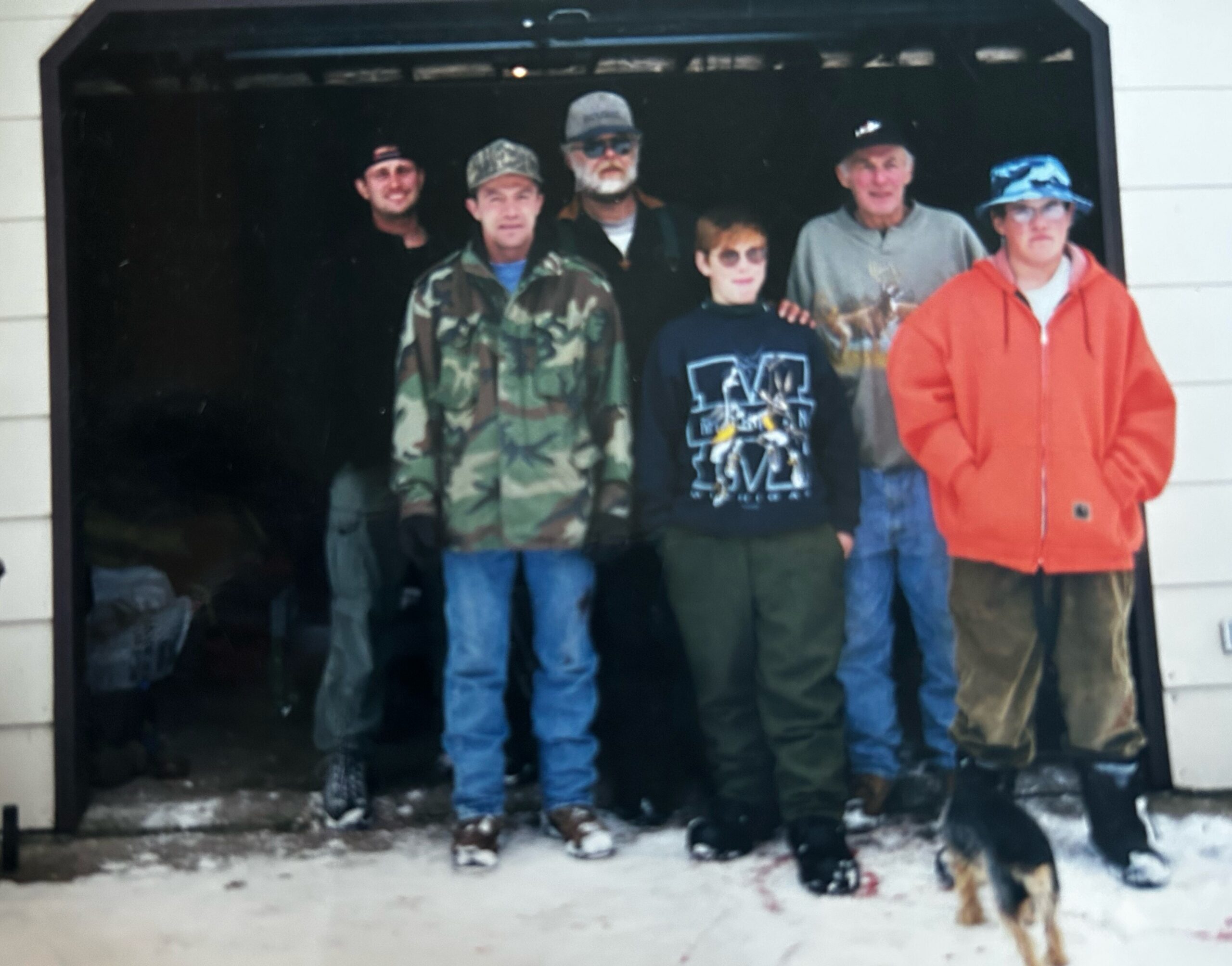 Six adults stand together outdoors in winter clothing, with a dog at the snowy ground in front of them.