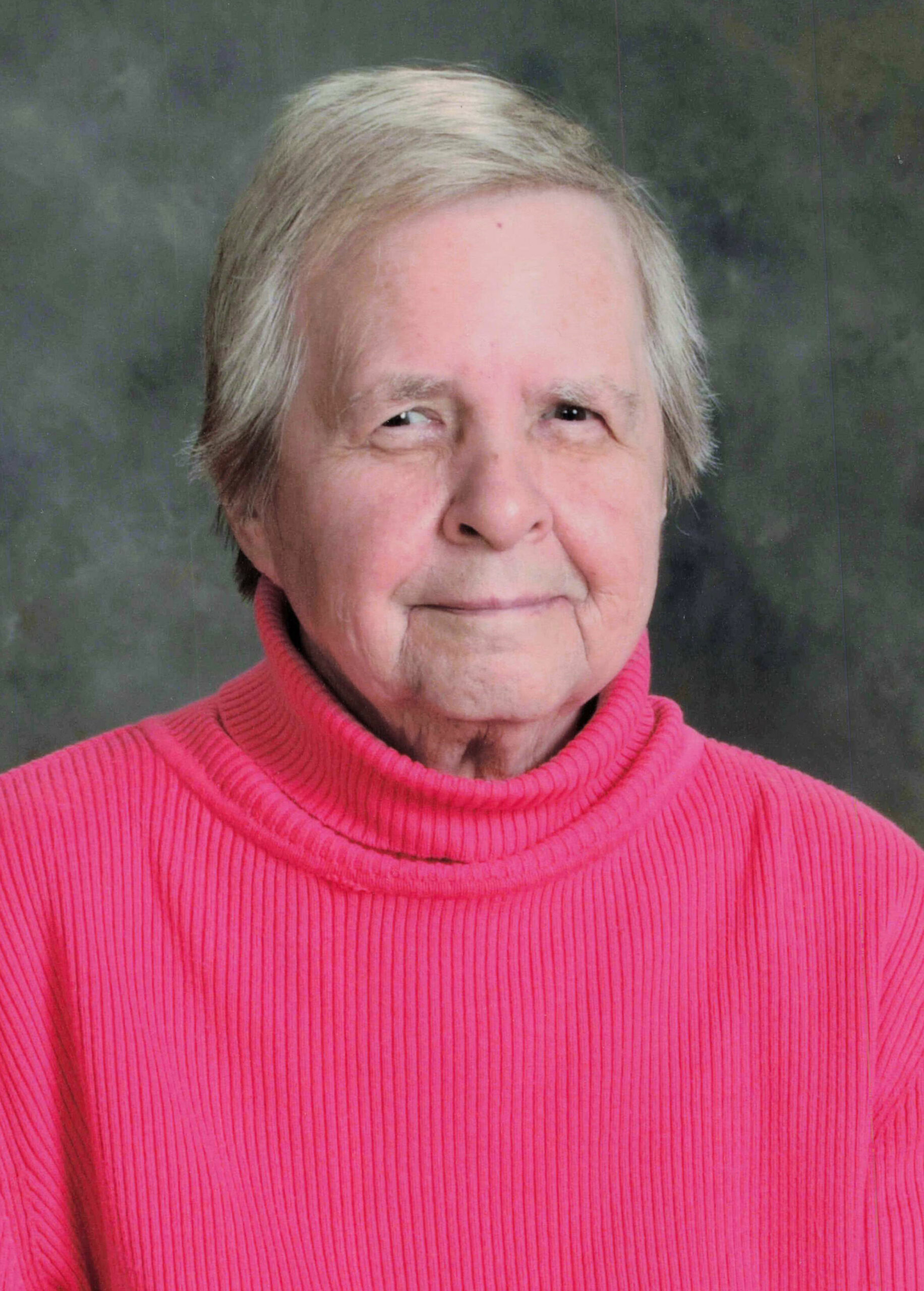 Close-up portrait of an elderly woman with short gray hair wearing a bright pink turtleneck sweater against a neutral backdrop.