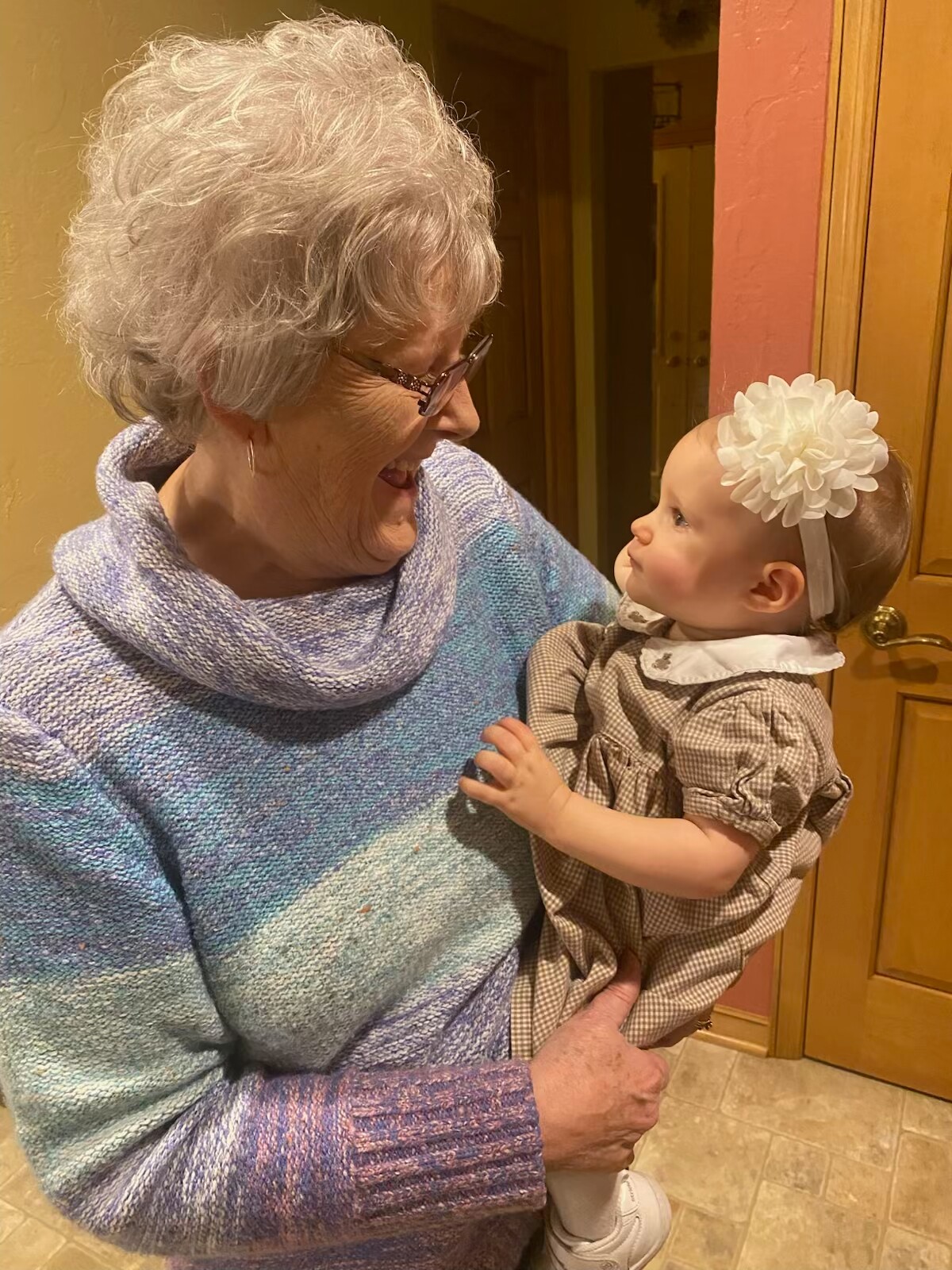 Smiling grandmother in a blue knit sweater holds a baby girl with a white flower headband, indoors.