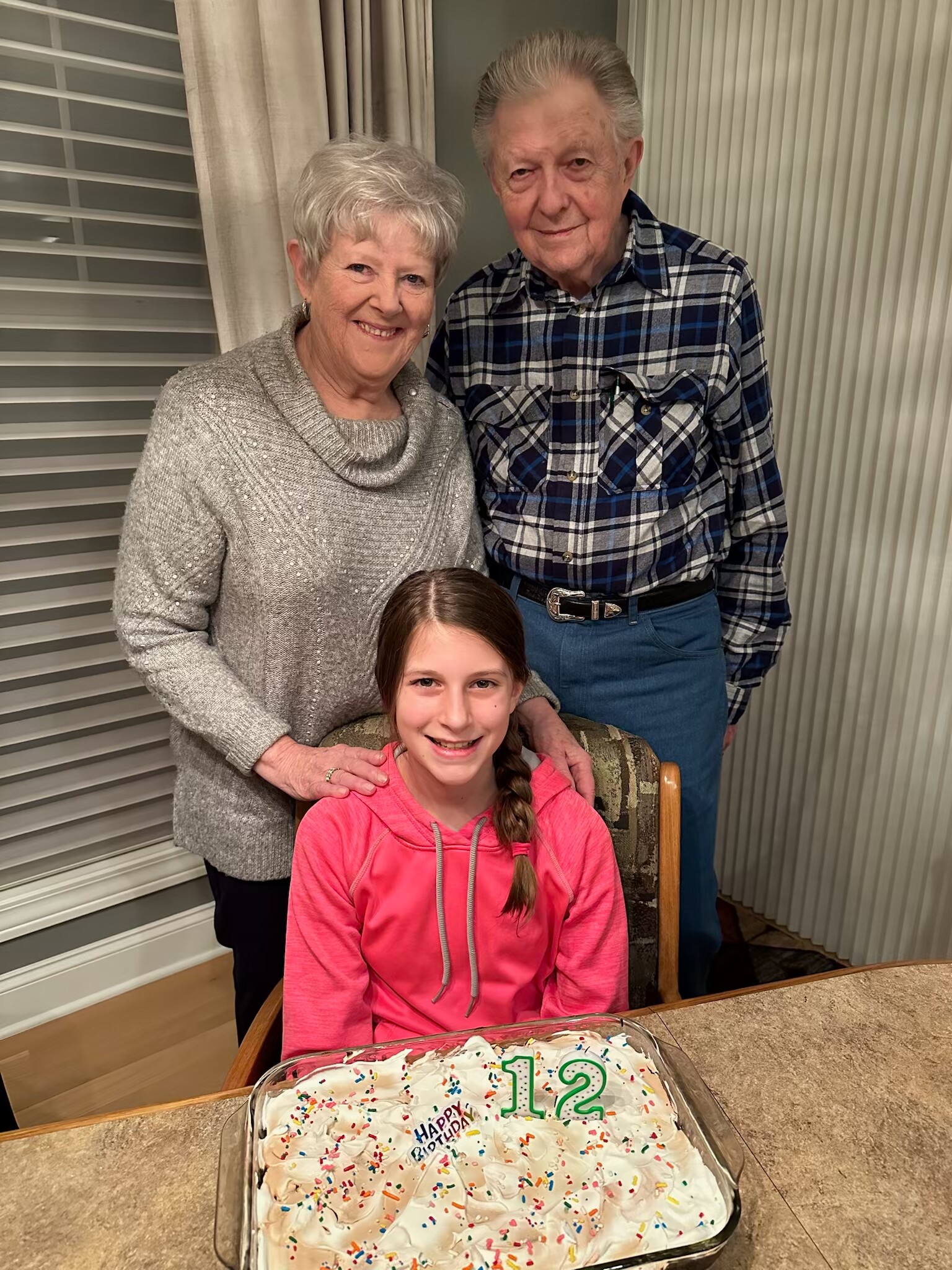 Smiling elderly couple and a girl pose with a birthday cake displaying the number 12 on a kitchen counter.