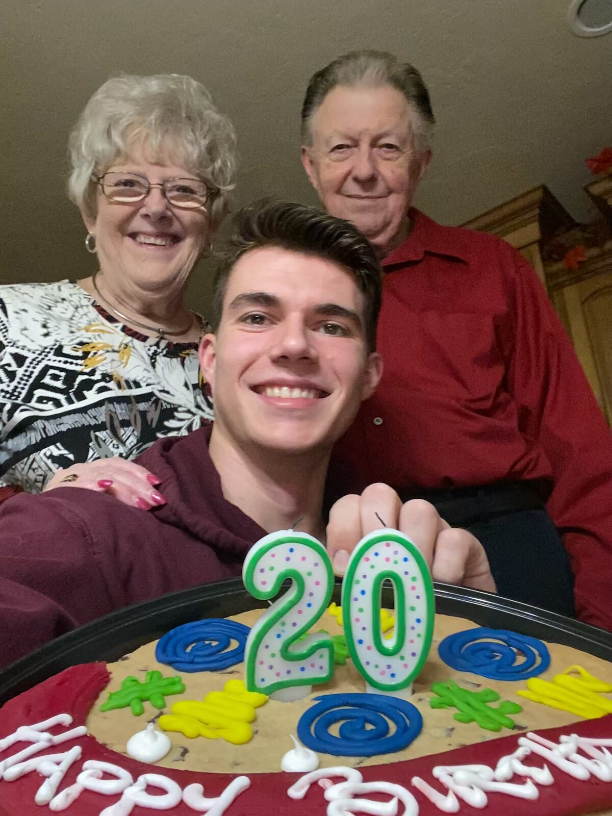 Young man taking a selfie with an older couple behind him at a birthday party; cake shows 20 candles.