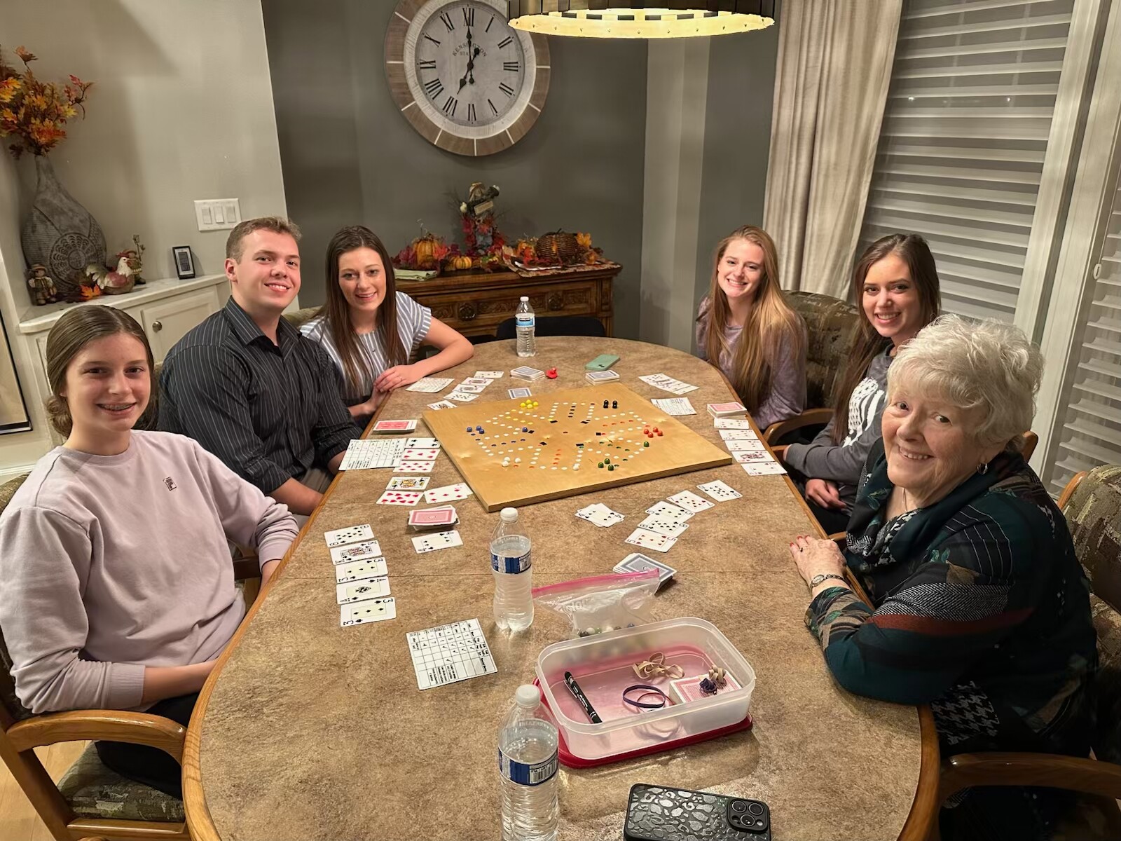 Six people around a kitchen table playing a board game, cards and small tokens spread across the table, everyone smiling at the camera.