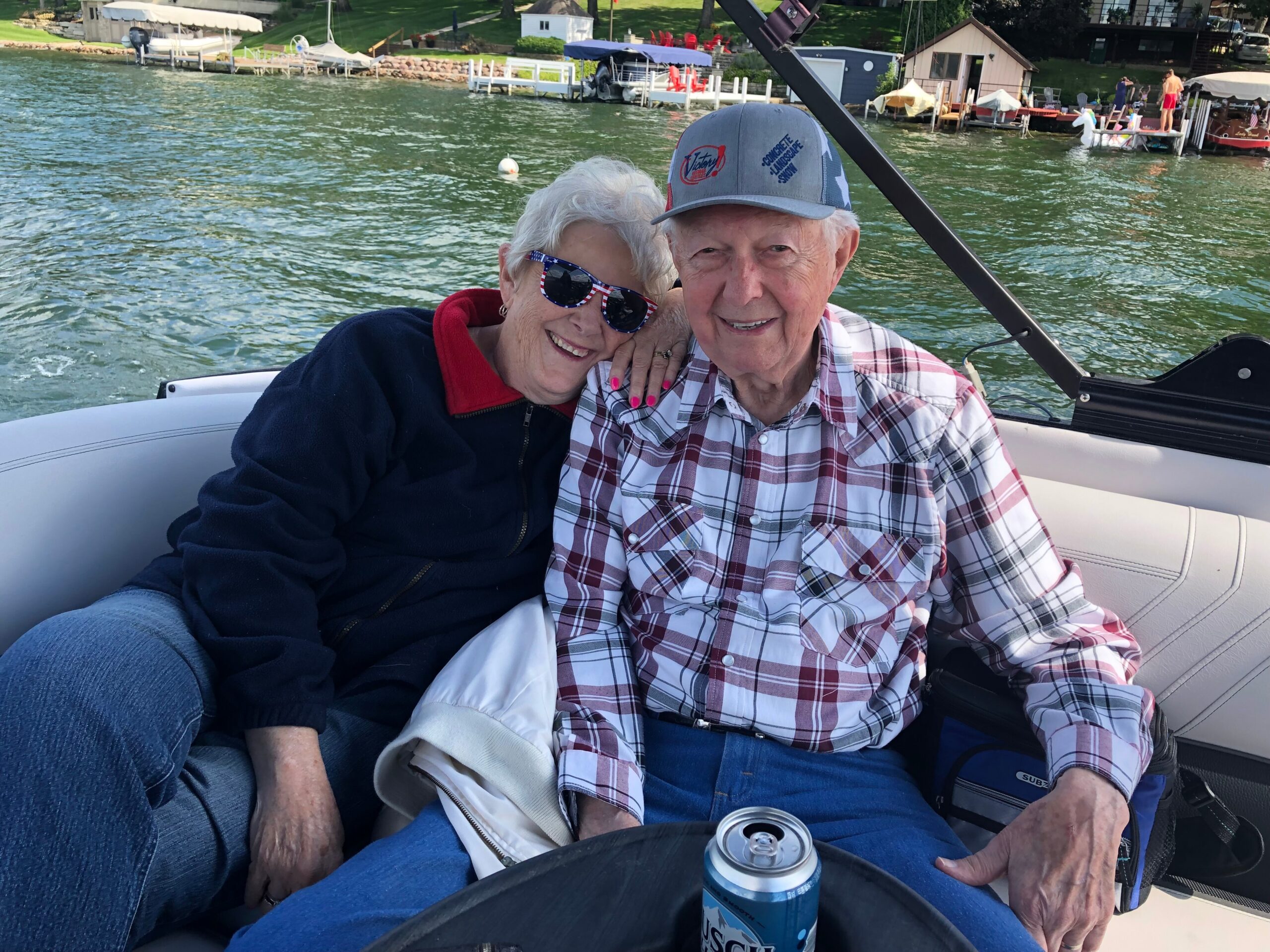 Smiling elderly couple sits close together on a boat with the lake and docks in the background.