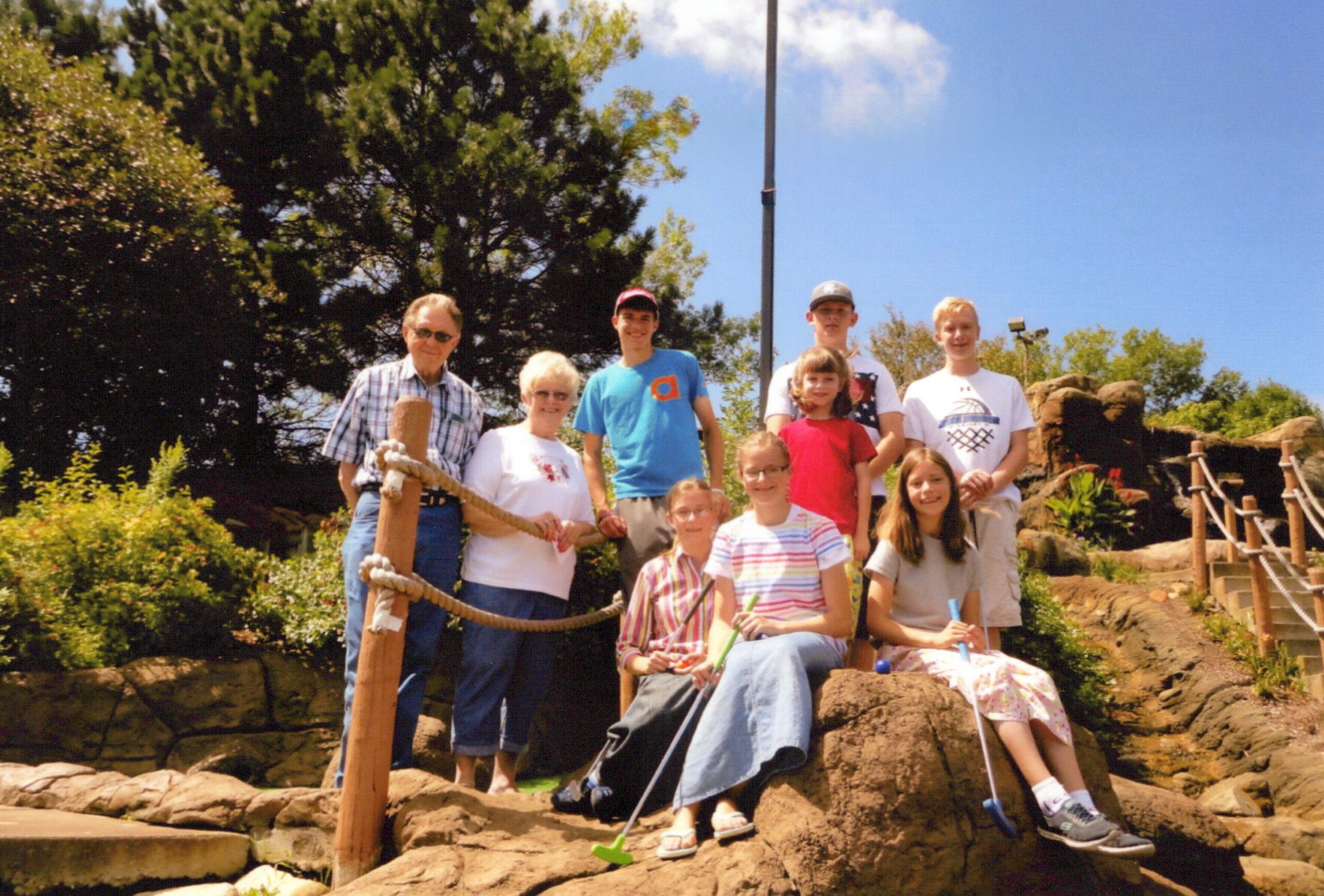 Group of people of varying ages posing together on a rocky outdoor play area with ropes and railings under a blue sky on a sunny day.