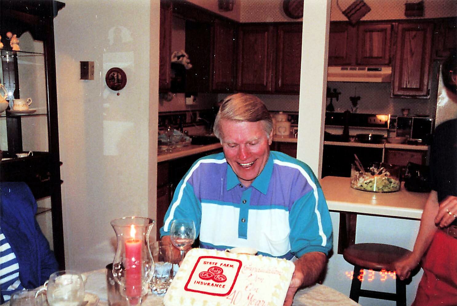 Smiling man in a blue striped polo at a birthday party, cutting a cake labeled with the State Farm Insurance logo.