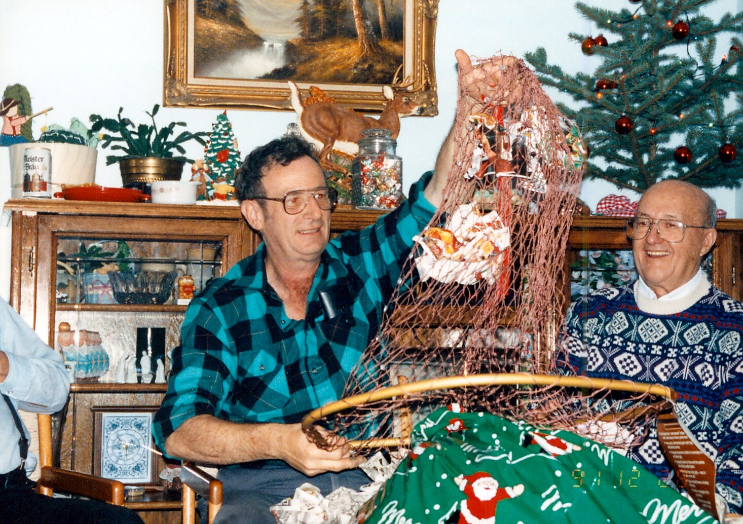 Two men celebrate Christmas at home, holding up a netted gift with wrapping paper nearby a decorated tree.