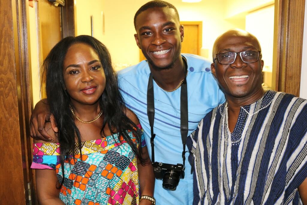 Three friends pose together in a doorway: a woman in a colorful patterned dress on the left, a man in a light blue shirt with a camera around his neck in the middle, and a man in a blue striped tunic on the right, all smiling.