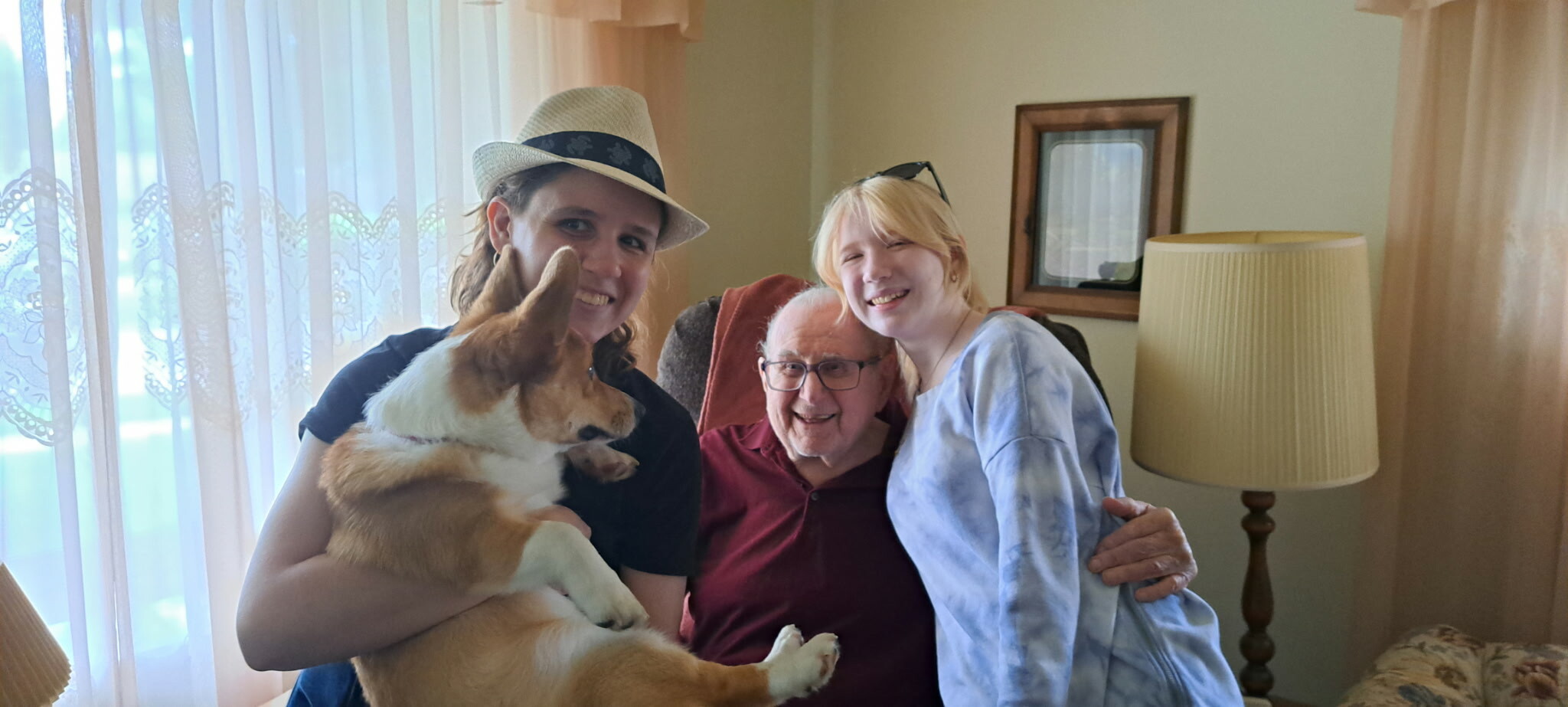 Woman wearing a straw hat holds a corgi while two others smile beside her in a living room setting.